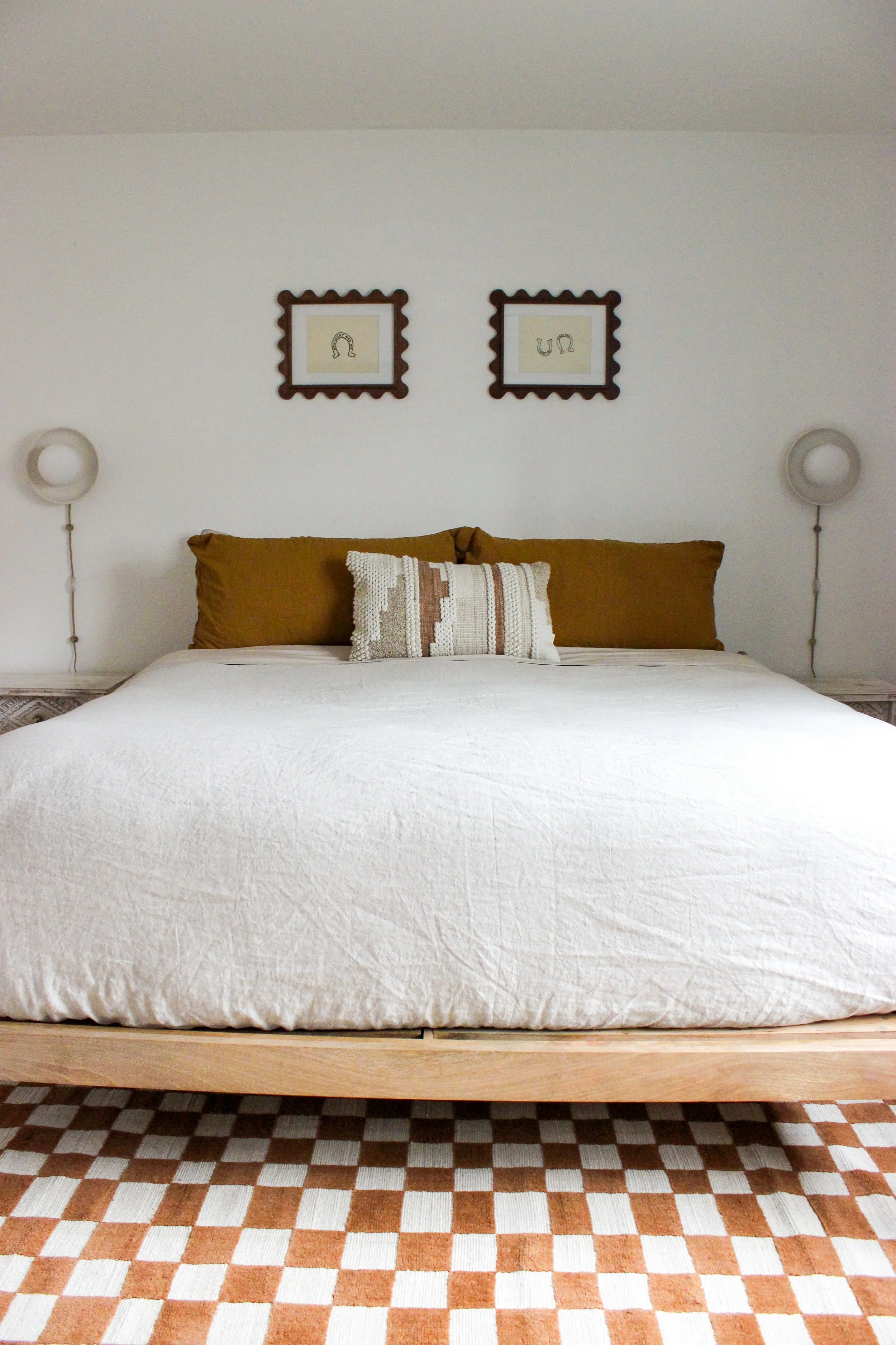 A cozy bedroom with a bed featuring a white quilt, four pillows with brown and beige covers, and a decorative beige, brown, and white pillow in front. Two round wall-mounted lamps are on either side of the bed, and above it are two framed pictures with scalloped borders and abstract line drawings, hung on a plain white wall. The room has a chevron-patterned rug with neutral tones.