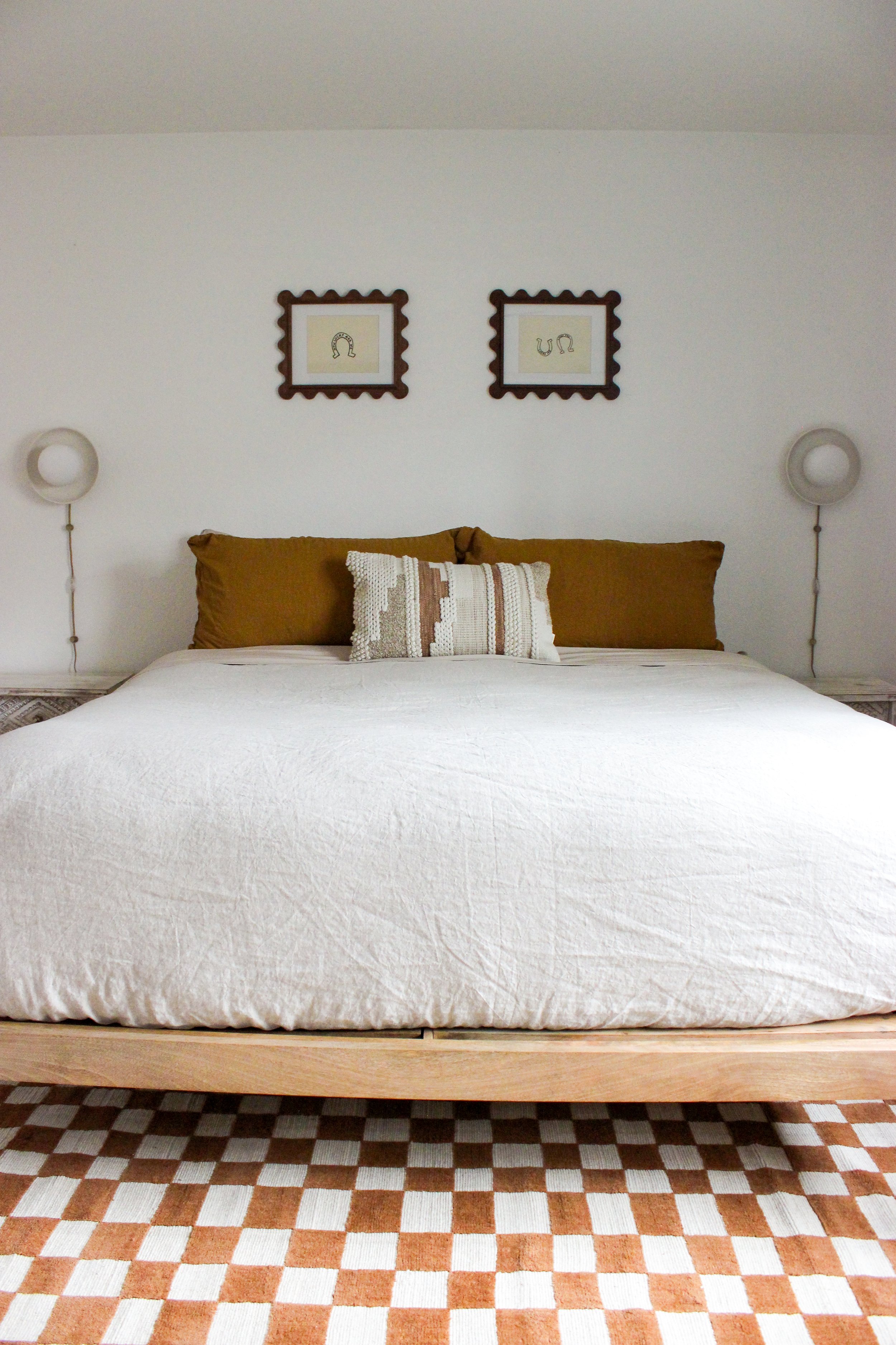 A bed with white bedding, two brown pillows, and a decorative pillow in the center, against a plain white wall, with two round white wall lamps on either side, and framed artwork above featuring horseshoes, on a checkered brown and white carpet.