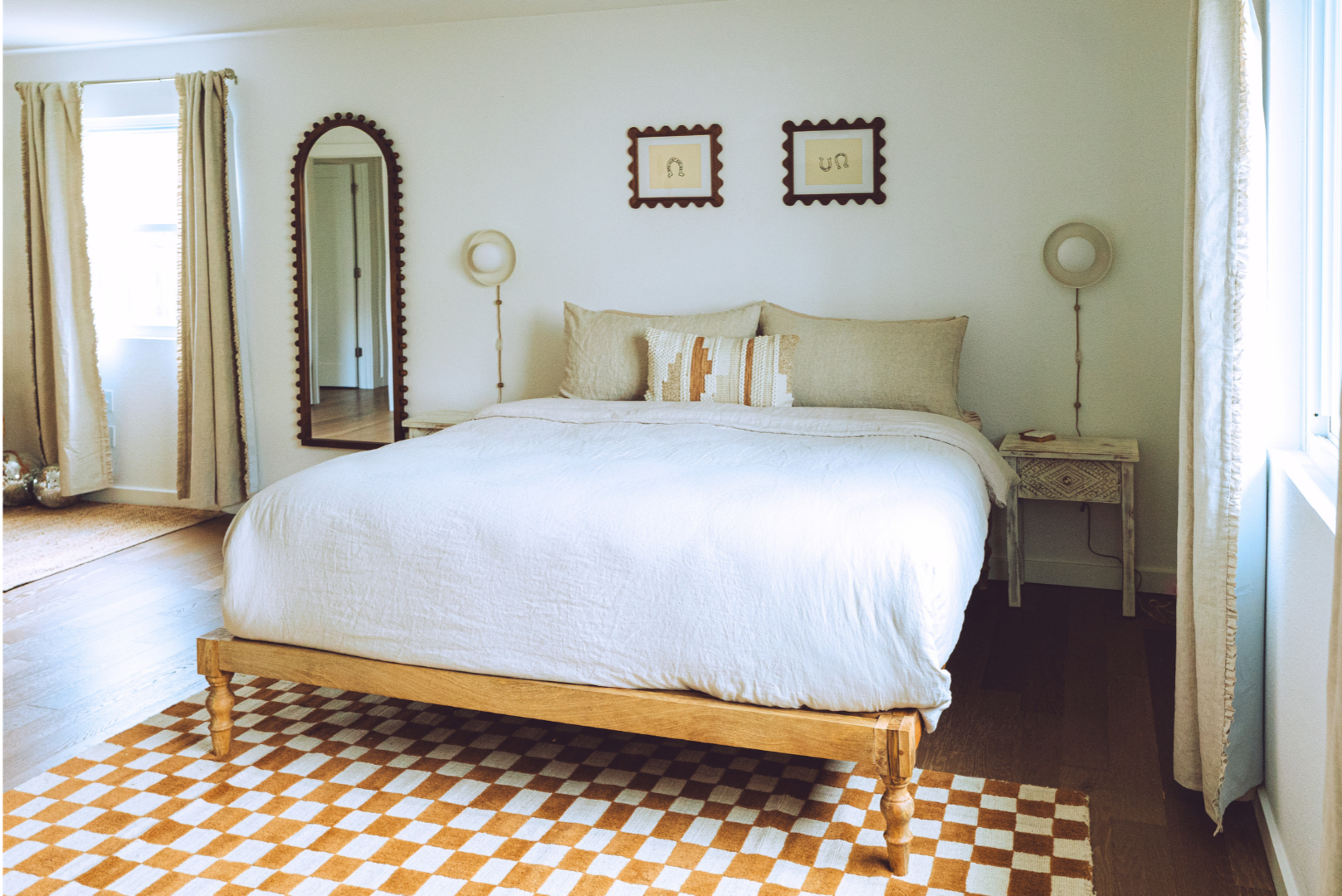 A bedroom with a white bed, beige pillows, and a patterned throw pillow. Nightstands with wall-mounted lamps on each side. A mirror and two framed artworks above the bed. Windows with beige curtains and a checkered orange and white rug.