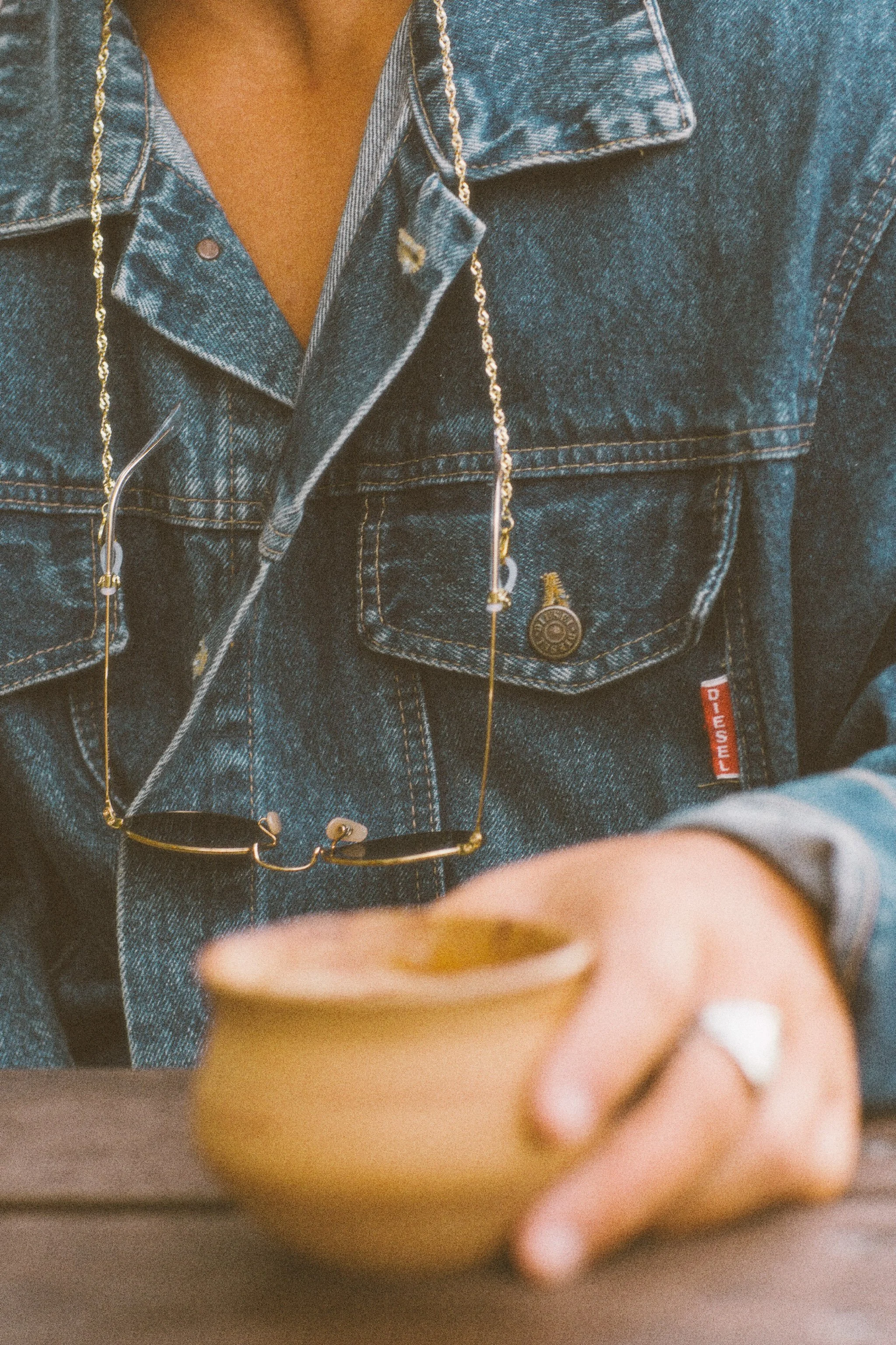 A person wearing a blue denim jacket with sunglasses hanging from the collar, holding a small beige cup.
