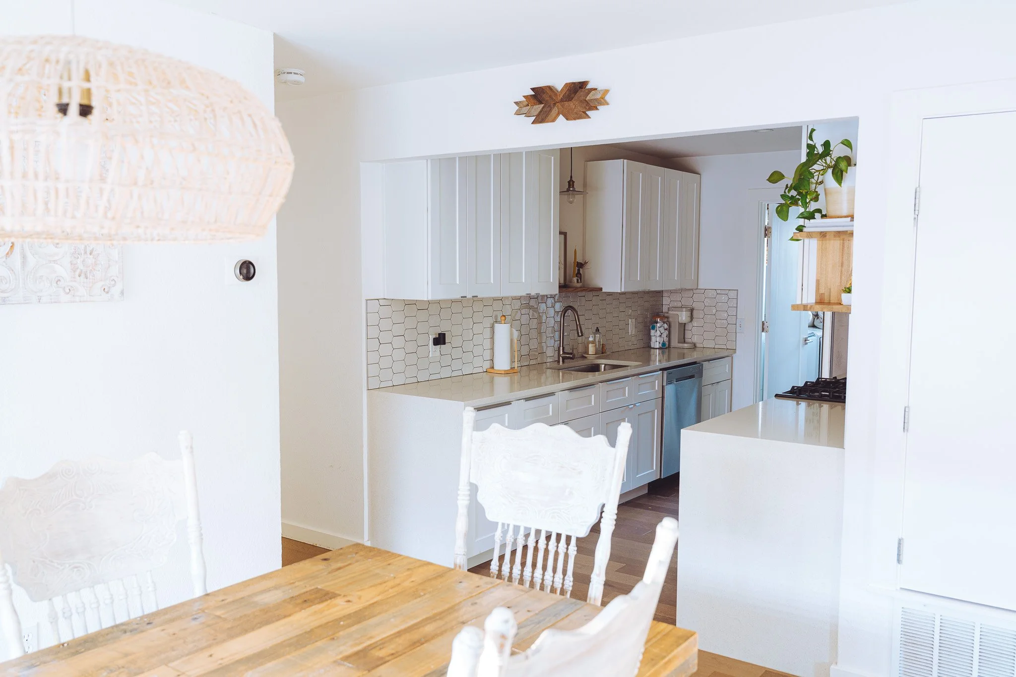 A kitchen with white cabinets, a hexagonal tile backsplash, a coffee machine, a plant on a wooden shelf, and a wooden dining table with white chairs.