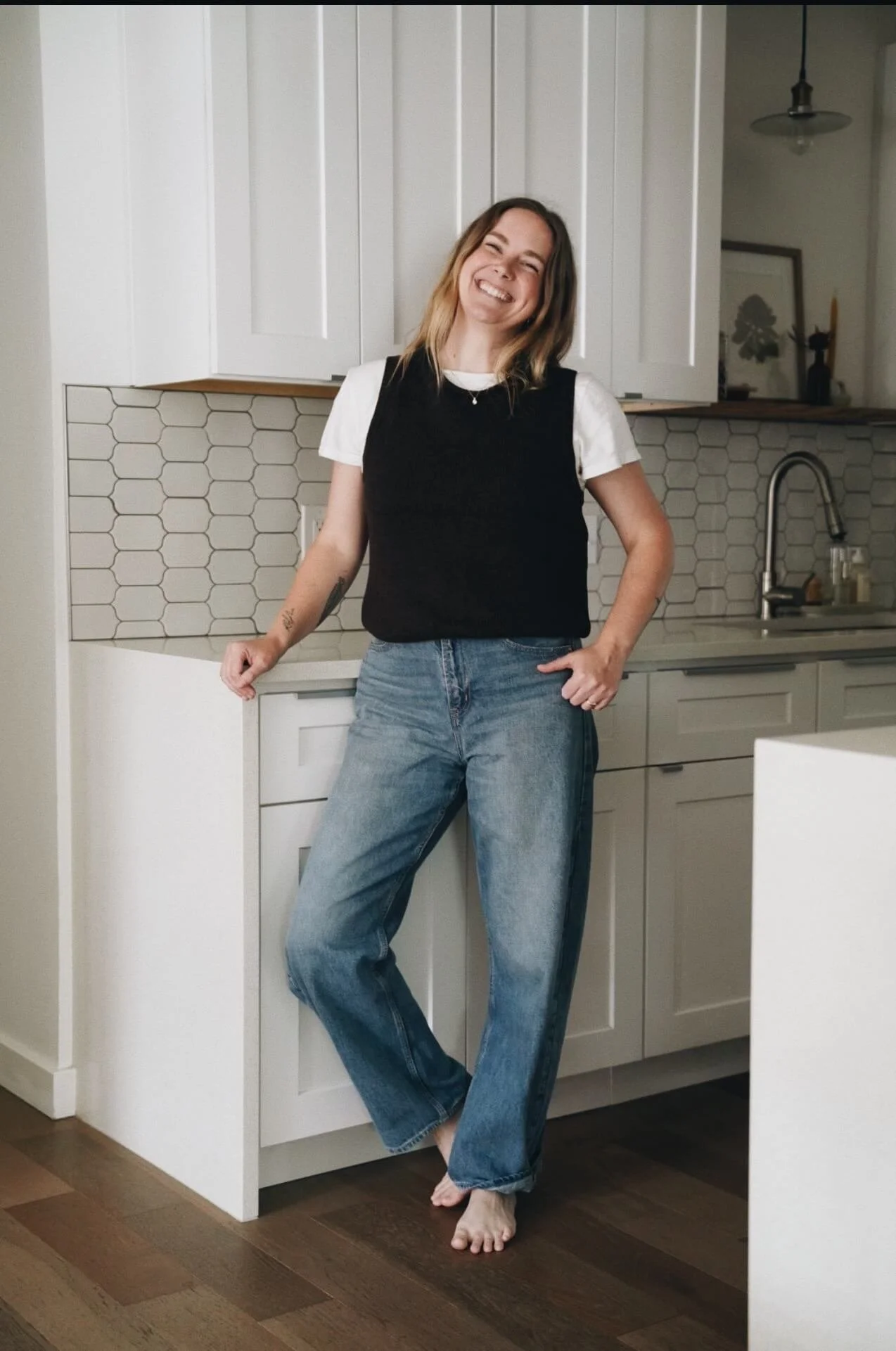 A woman with shoulder-length blonde hair smiling and standing barefoot in a kitchen, wearing a white t-shirt, black vest, and blue jeans, leaning against a white kitchen counter with hexagonal tile backsplash.