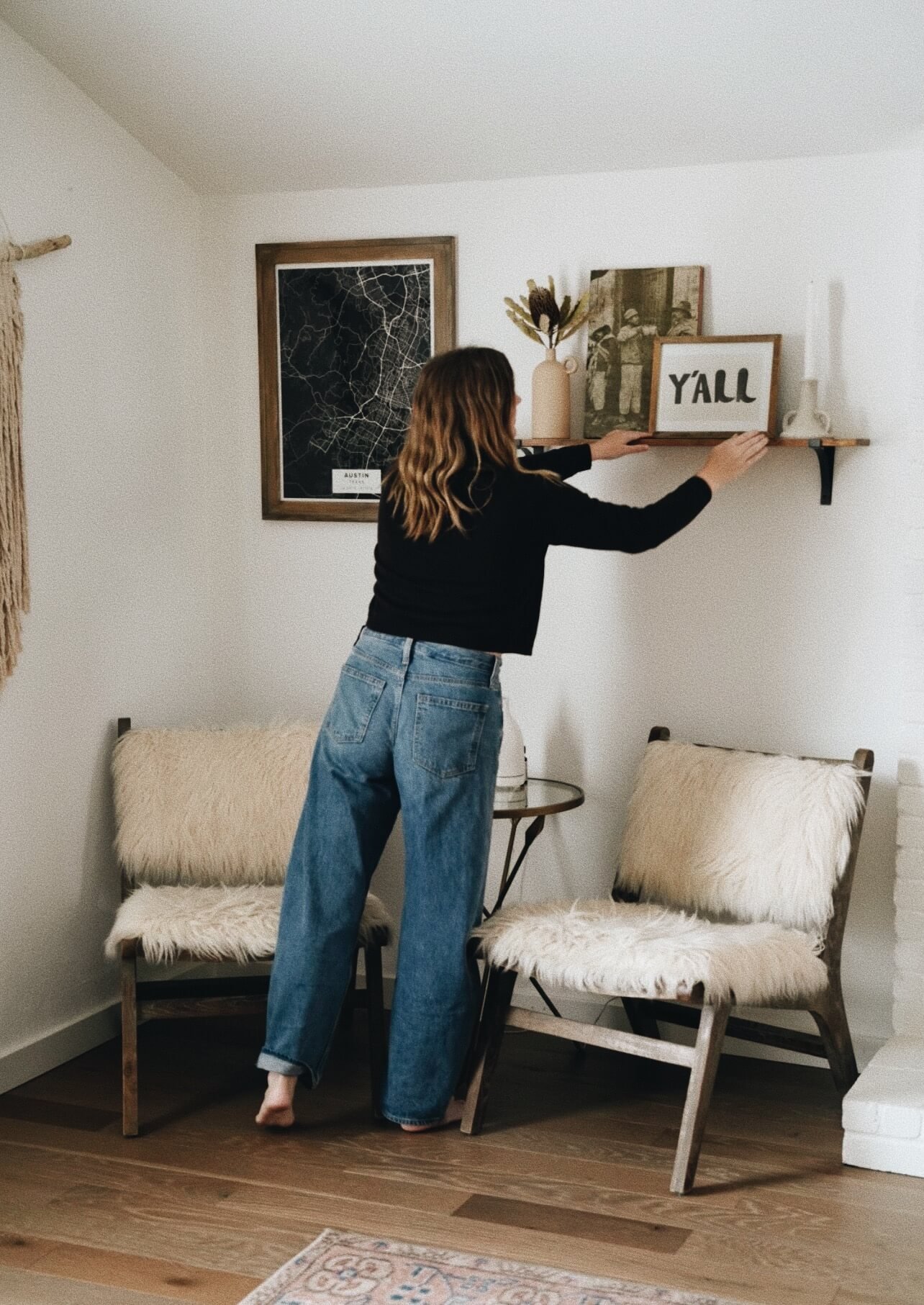 A woman with wavy hair, dressed in a black top and blue jeans, arranging decor on a wall shelf in a living room with two cushioned chairs and wall art.
