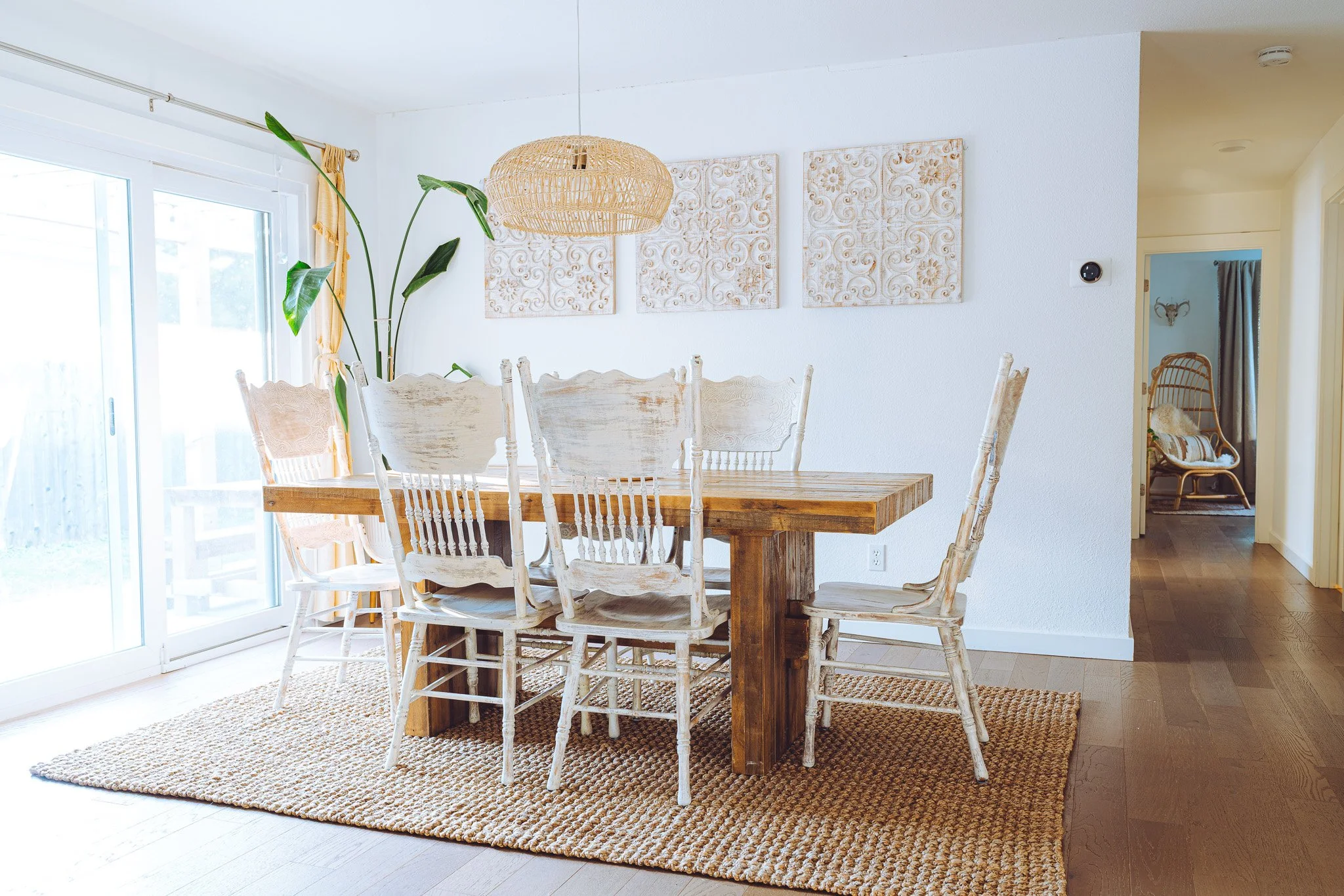 A dining room with a wooden table and white distressed chairs on a woven rug, large windows with yellow curtains, a plant, and wall art, with a hallway visible in the background.