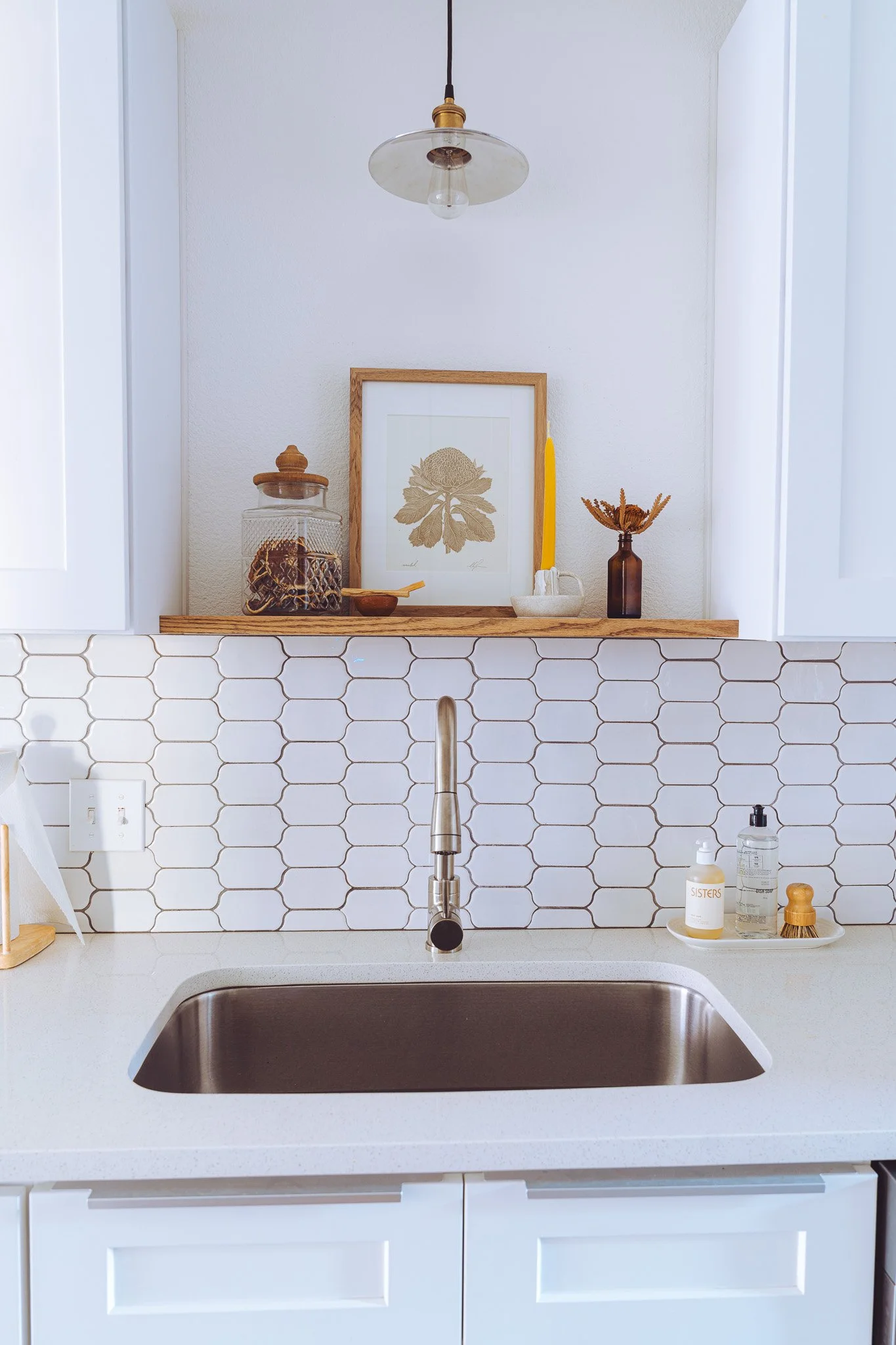 Close-up view of a kitchen sink with a modern brushed metal faucet, white countertop, and a white tile backsplash with a honeycomb pattern. Above the sink, there is a wooden shelf holding decorative items including a framed botanical print, a clear j