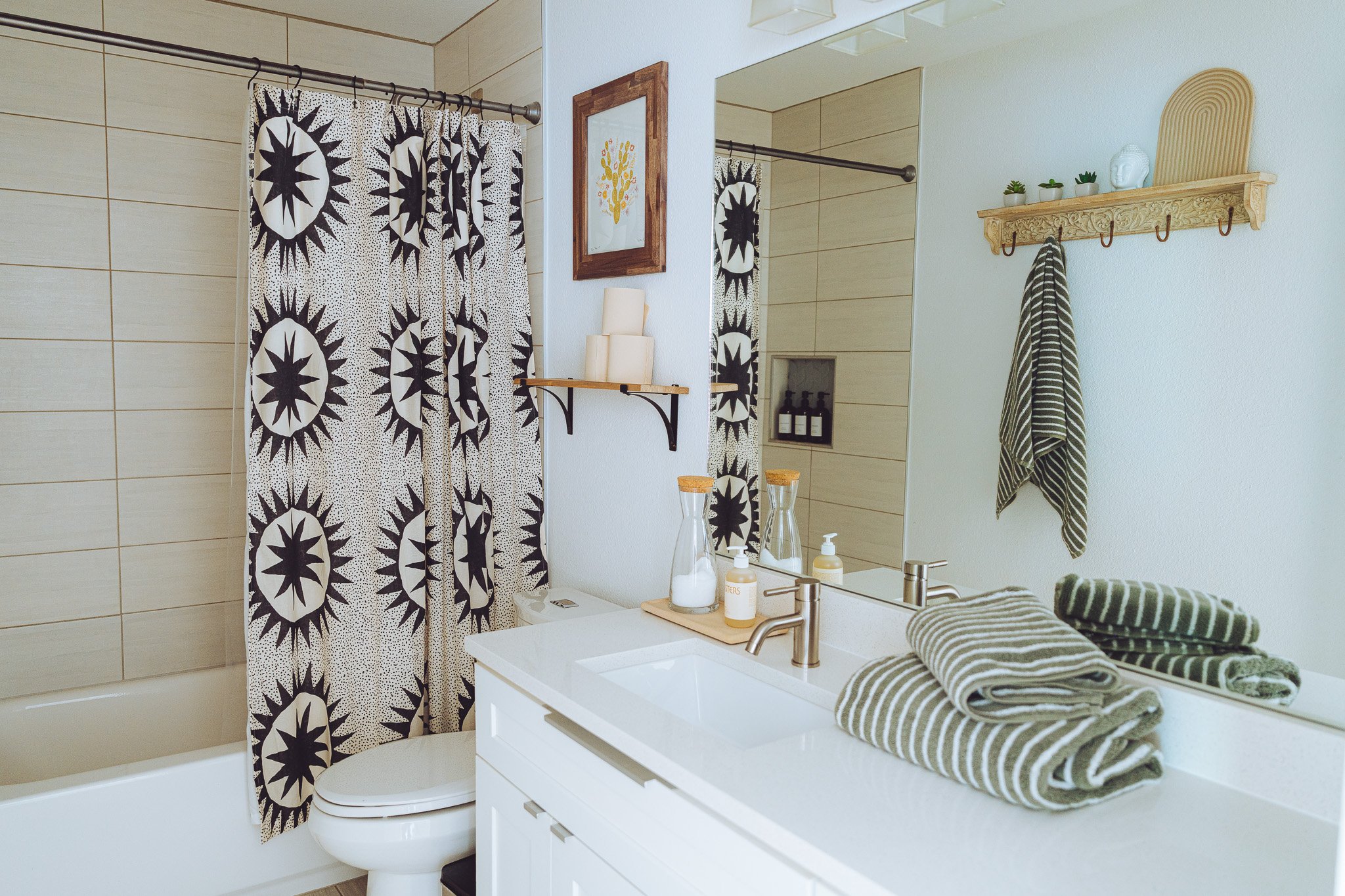 A bathroom featuring a vanity with a white countertop, a large mirror, and neatly folded striped towels. Next to the vanity, a shelf with miniature potted plants and decorative items is mounted on the wall. A shower area with a black-and-white patter