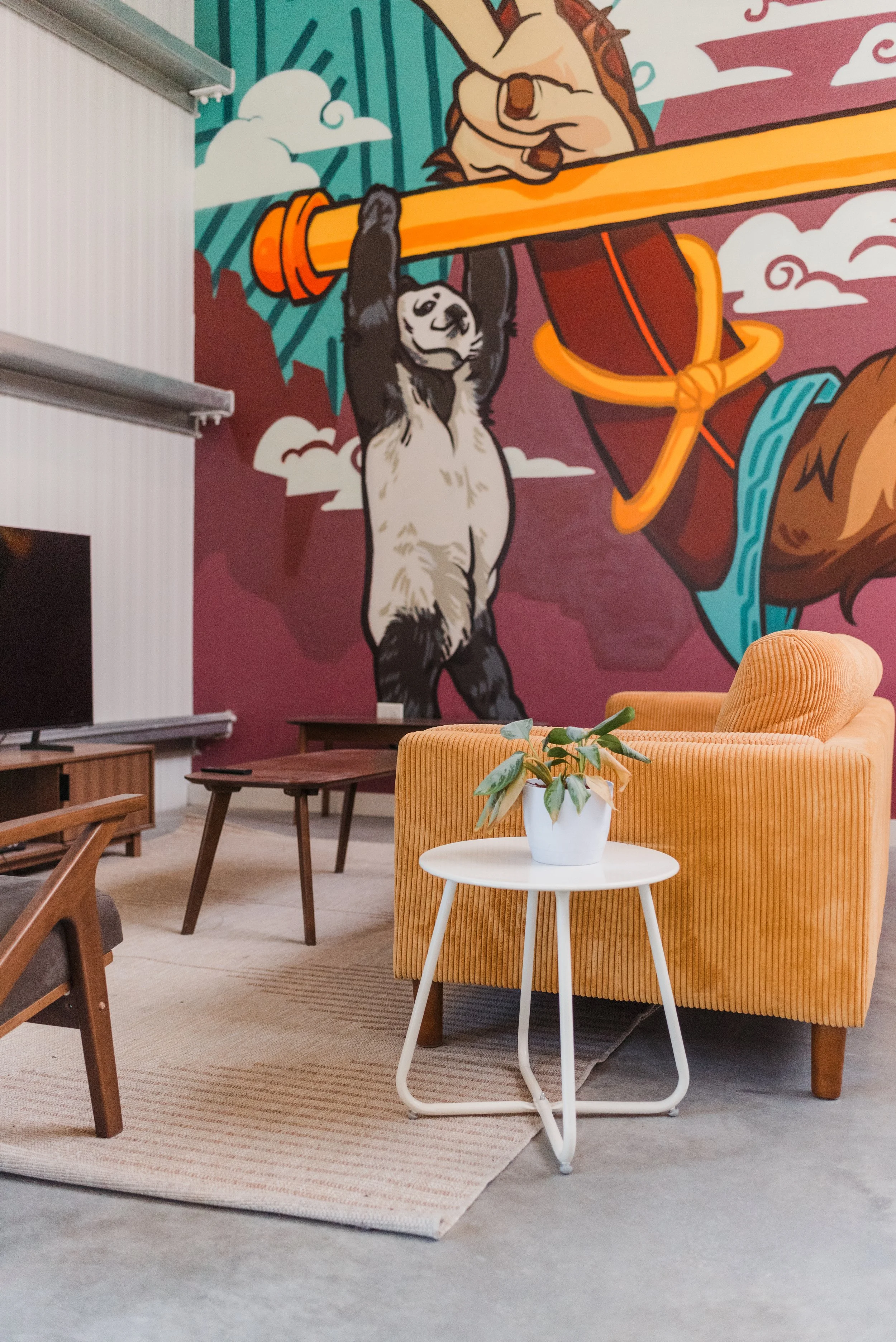 Cozy living room with a yellow upholstered armchair, a white side table with a potted plant, and a mural painting of a sloth hanging from a pole on the wall.