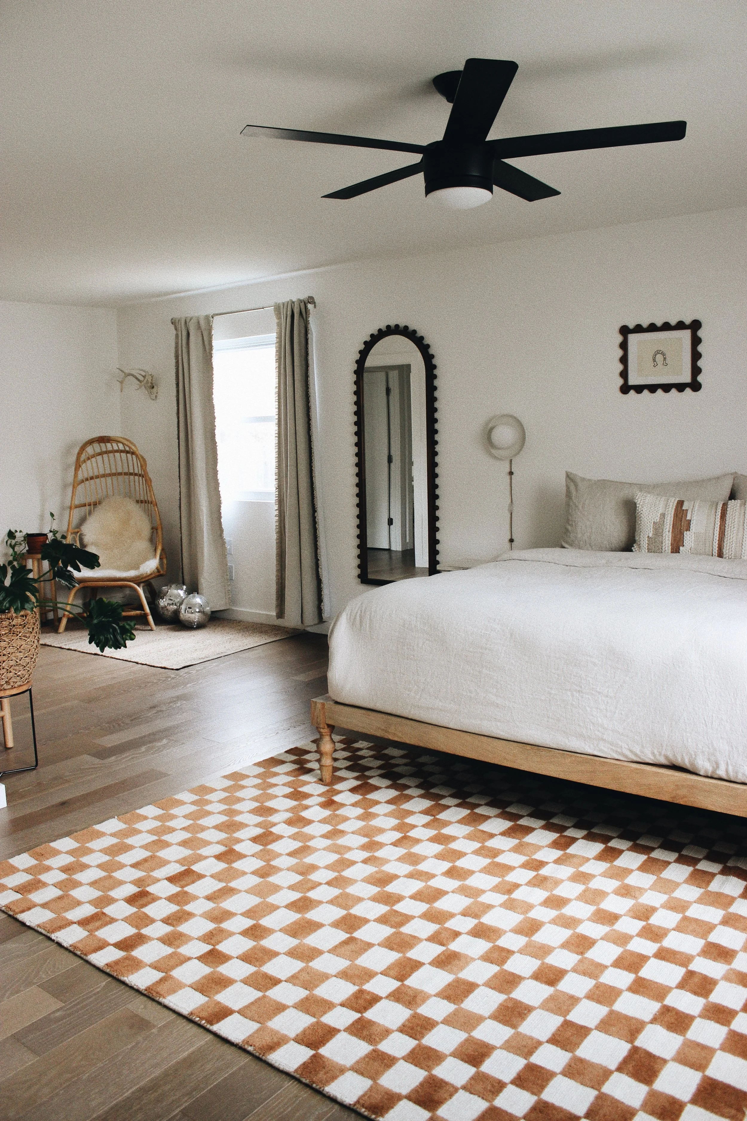 A bedroom featuring a large bed with white bedding and pillows, a patterned orange and white area rug, a mirror with a black scalloped frame, a bed-side wall lamp, a wicker chair with a white cushion, plants, a framed piece of artwork with a horsesho