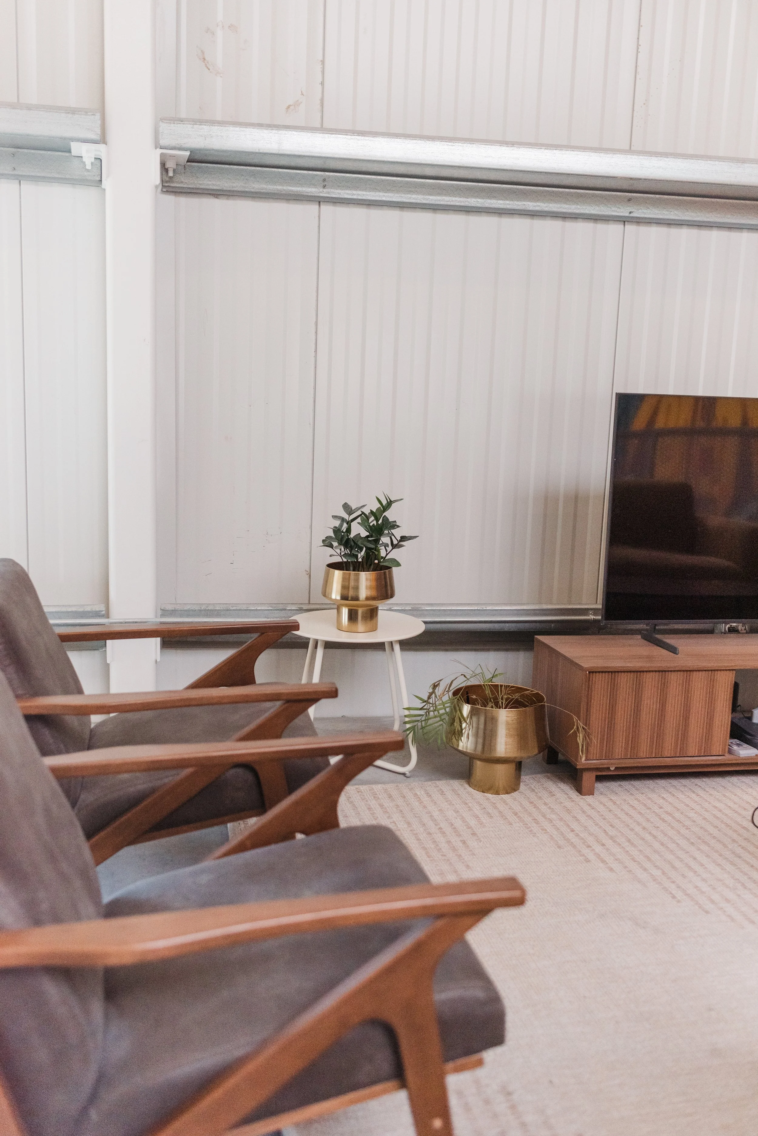A cozy living room corner with three gray armchairs with wooden arms, a small white side table with a potted plant in a gold planter, and a wooden TV stand with a flat-screen TV. There are two gold planters with plants near the table and TV stand. Th
