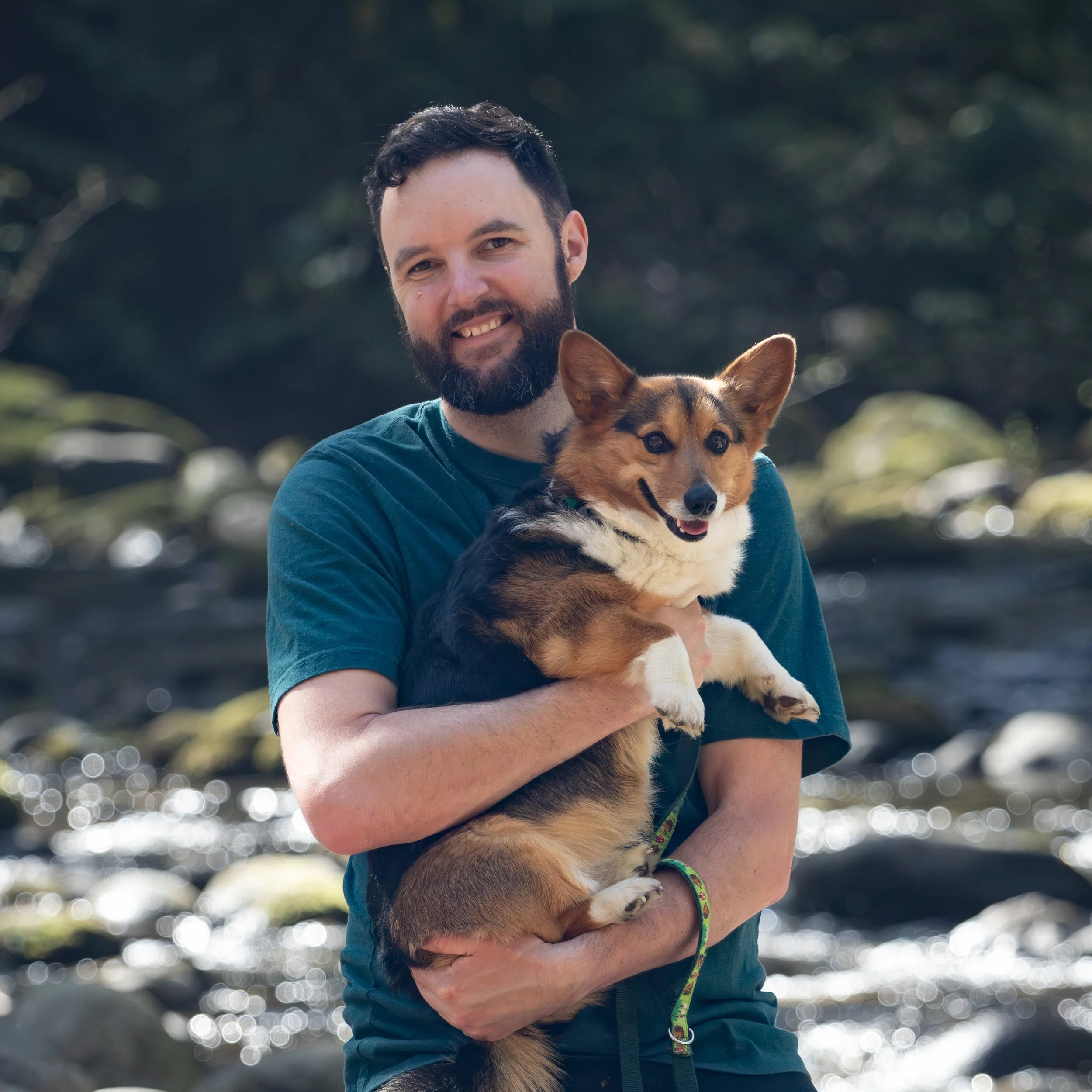 Jesse Heaslip, the owner and pack leader of Leaps and Bounds Pet Care is a tall man with a beard and dark hair smiling while holding a happy corgie with large ears and soft fur, outdoors near a rocky stream.