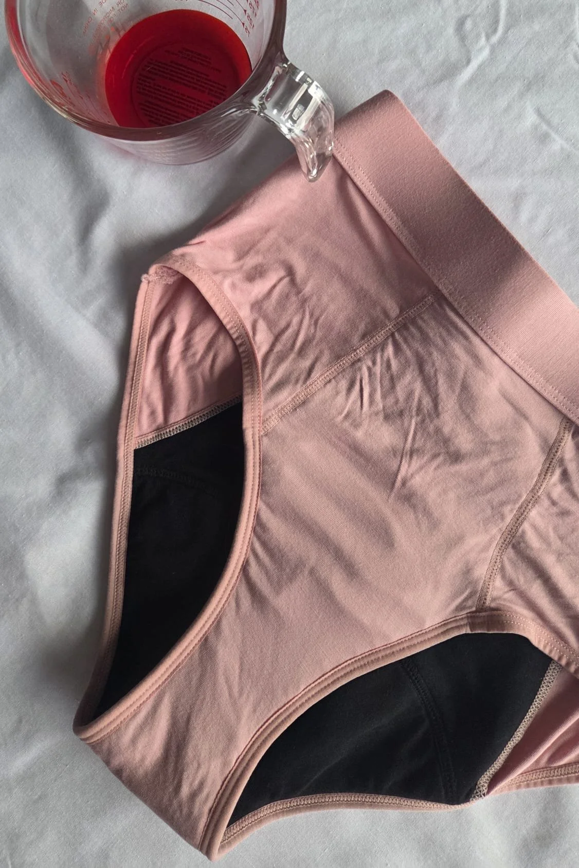 A pair of pink and black period undwear with a wide elastic waistband, placed on a white surface. A measuring cup with red liquid is positioned above the shorts.