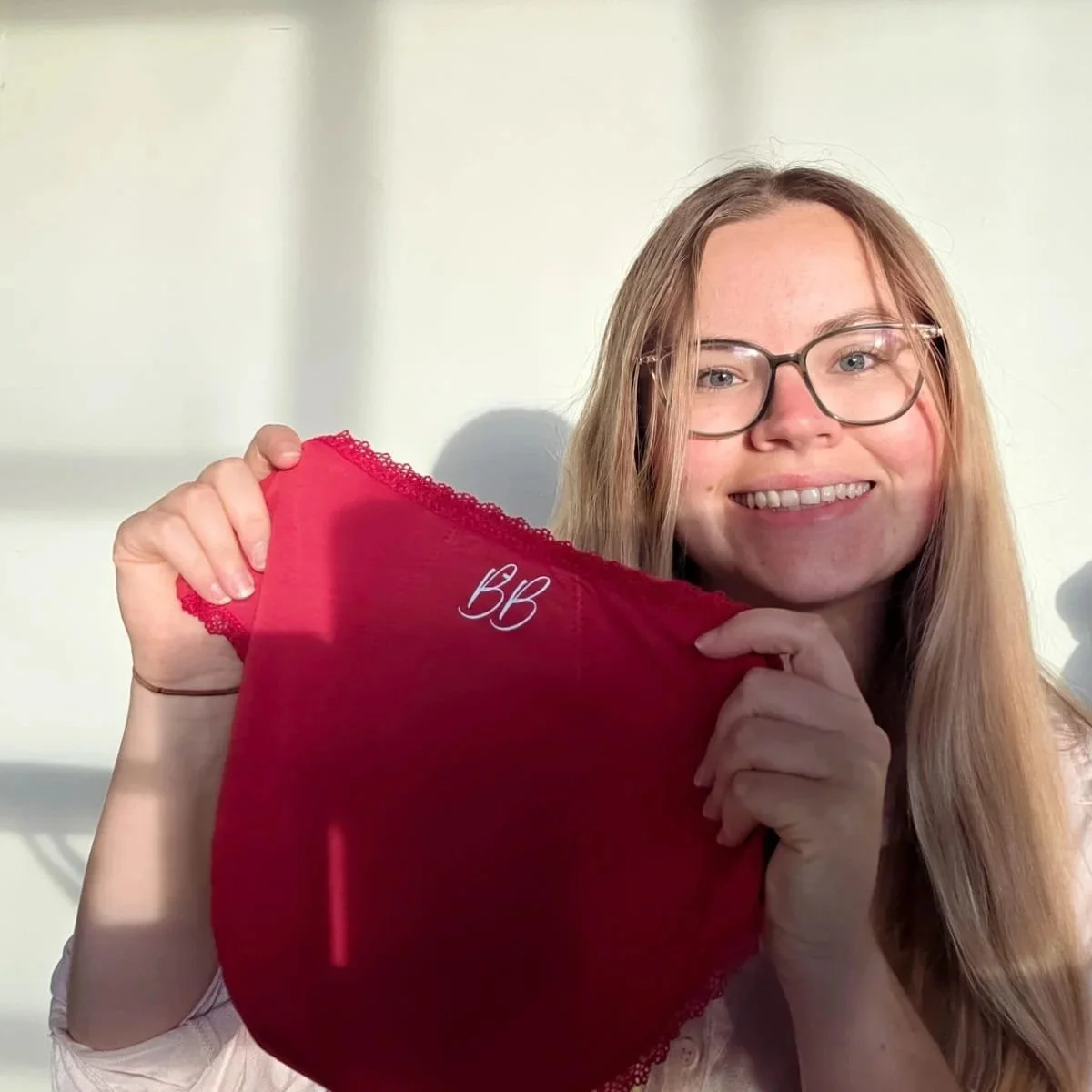 A smiling young woman with glasses holds a red piece of clothing with white embroidered initials 'BB' against a light-colored wall.