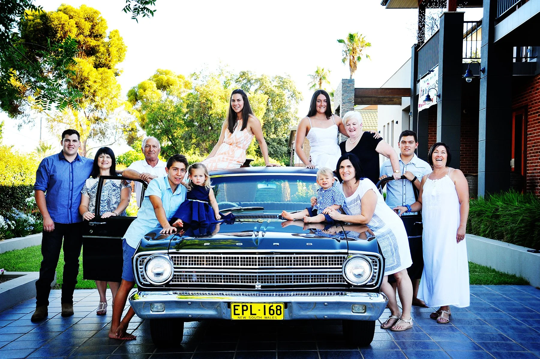A large group of people, including men, women, and children, gathered around and on a vintage black Ford car with a yellow New South Wales license plate, outdoors in a garden setting with trees and a building in the background.