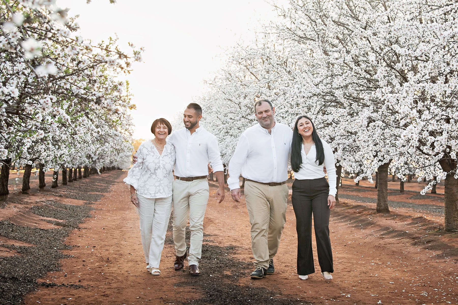 Four adults walking in a blossoming orchard of white flowers, smiling and enjoying the spring day. The group consists of two men and two women, dressed in casual and semi-formal clothing.