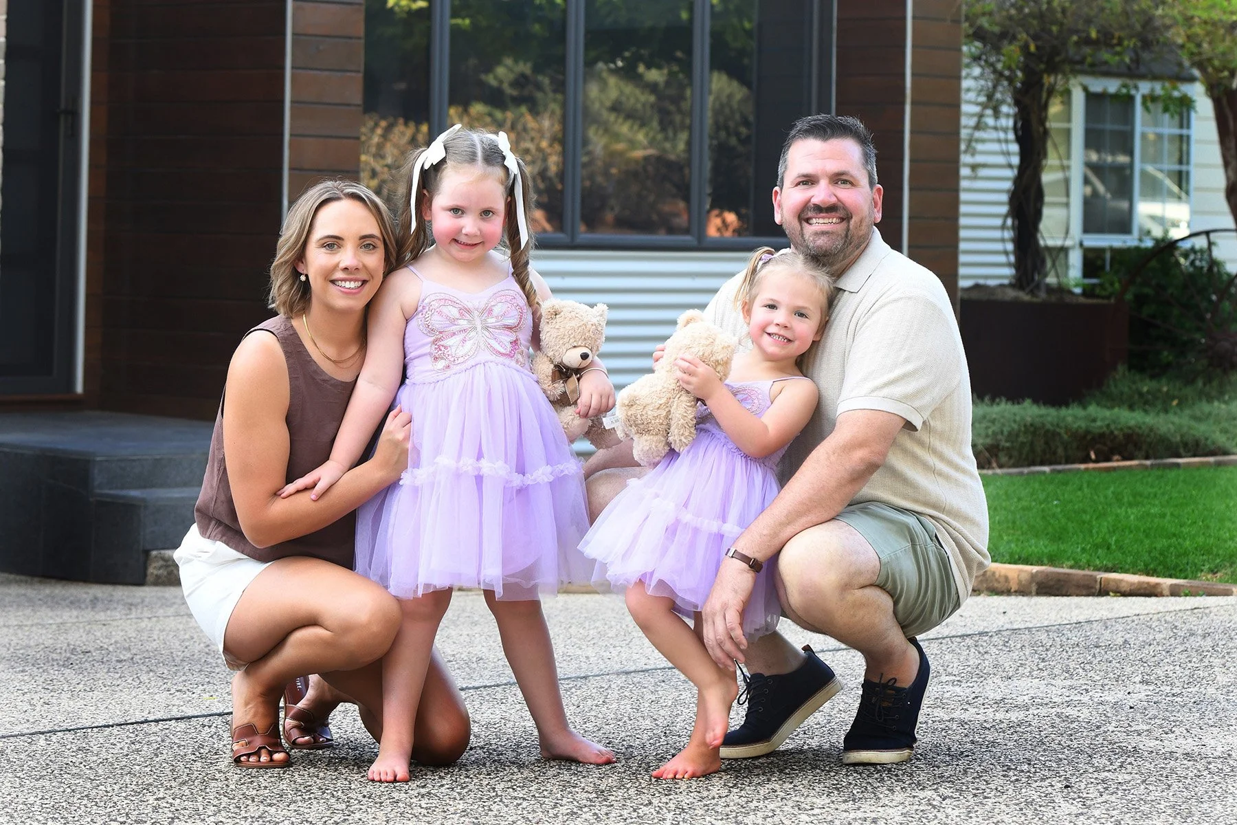 A family of five posing outdoors in front of a house with large windows. The mother and father are kneeling on the ground while two young girls stand between them, all smiling at the camera. The girls are wearing matching purple dresses and holding teddy bears. The family appears happy and relaxed.