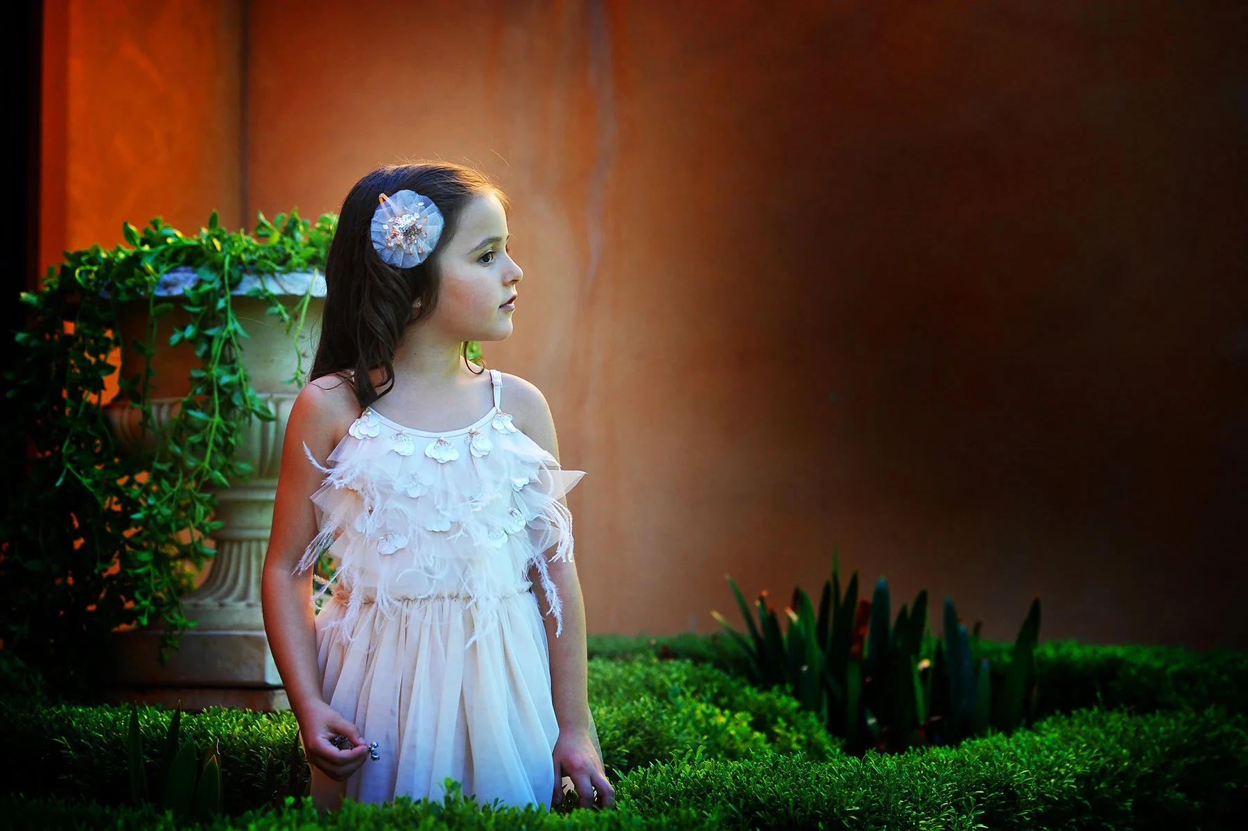 A young girl in a white dress with floral details standing outdoors near a large plant and greenery, gazing to her left.