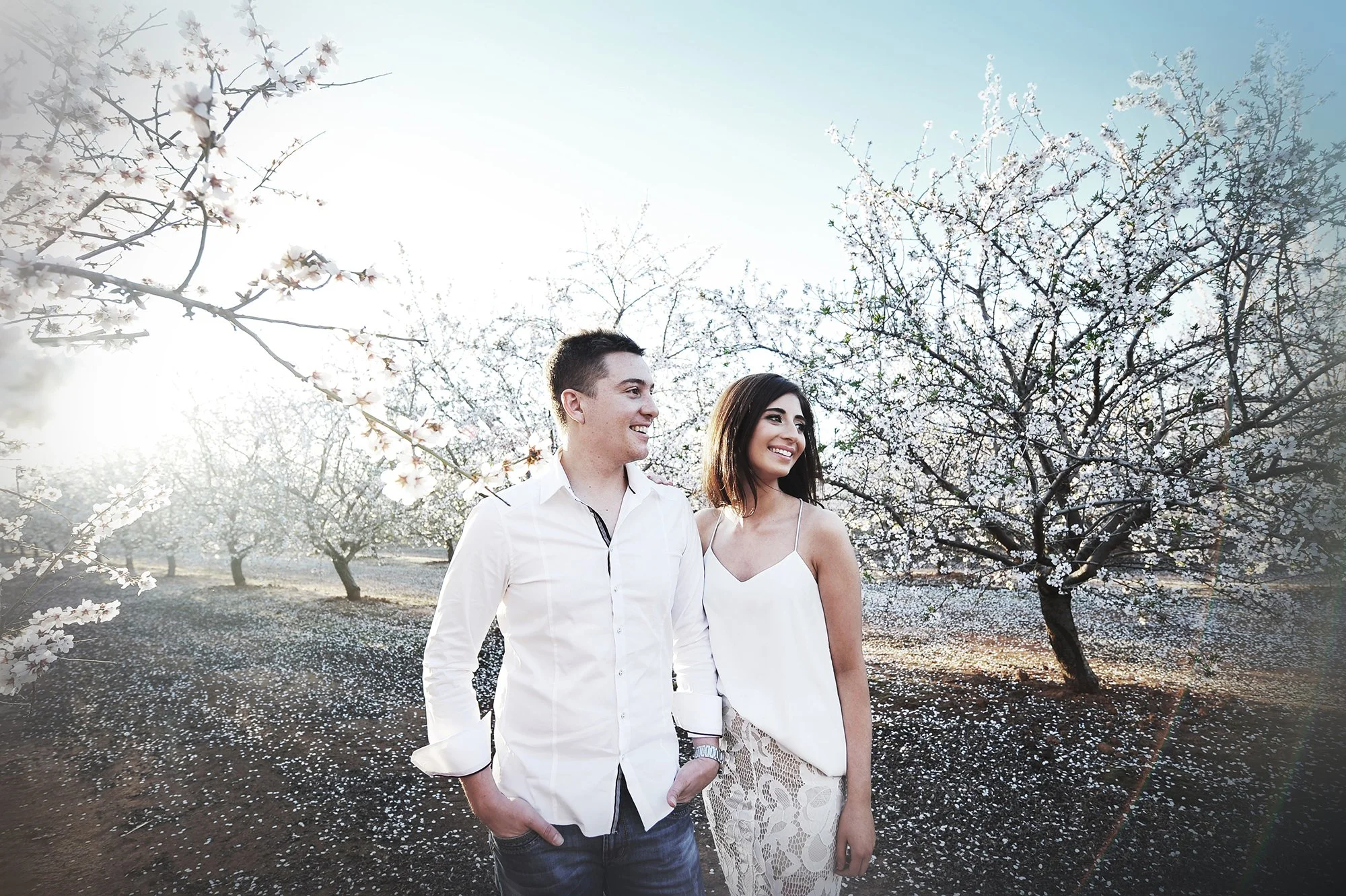 A young couple smiling and walking outdoors in a grove of blossoming trees during springtime, with sunlight filtering through the branches.