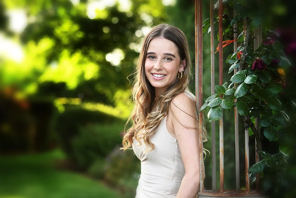 A young woman with long wavy hair, wearing a sleeveless white top and hoop earrings, is smiling and standing outdoors beside a rusty metal fence with greenery and flowers.