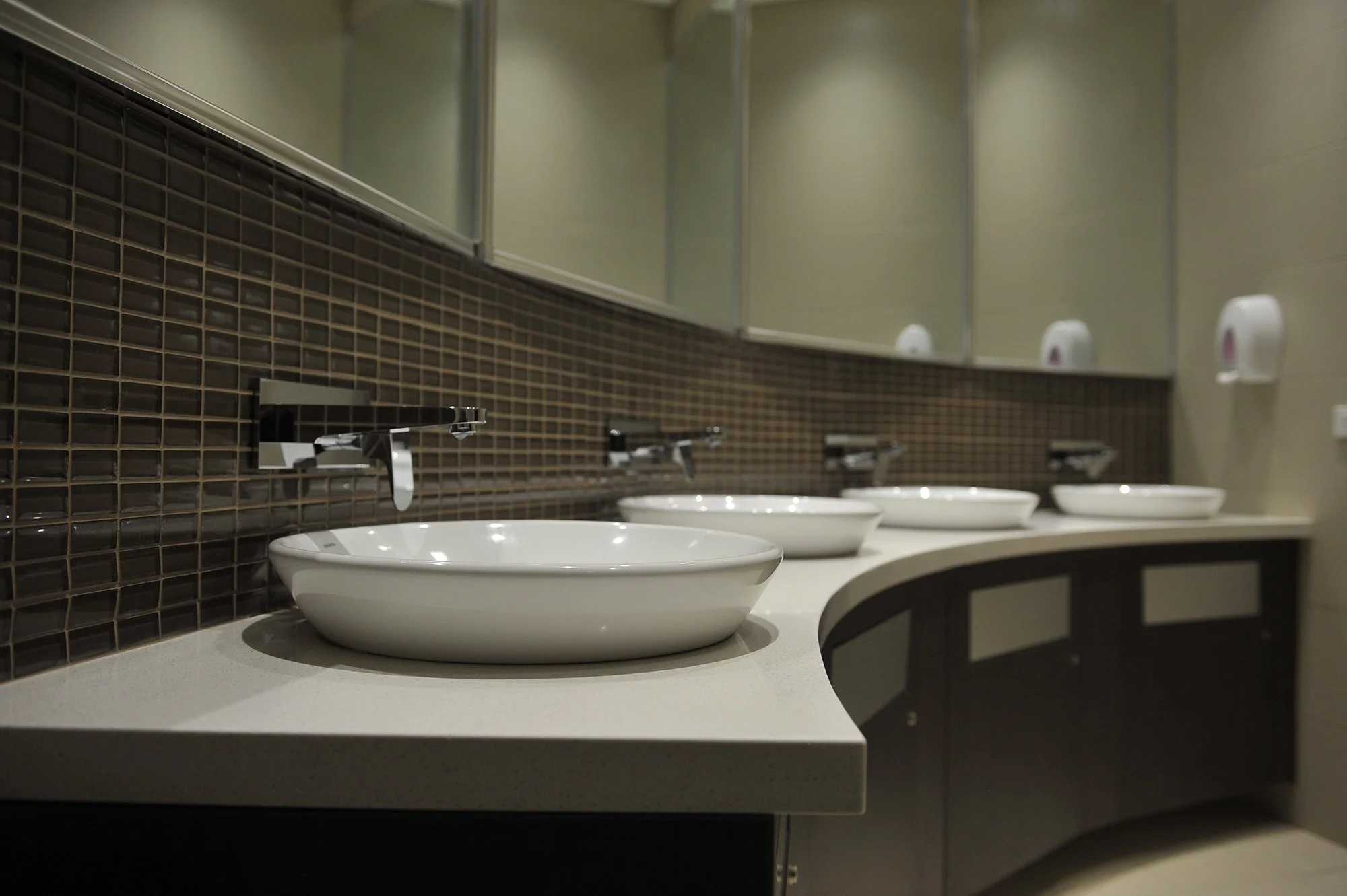 Multiple white ceramic sinks with chrome faucets in a public restroom, mirrored wall overhead, brown tile backsplash, soap dispensers on the wall.