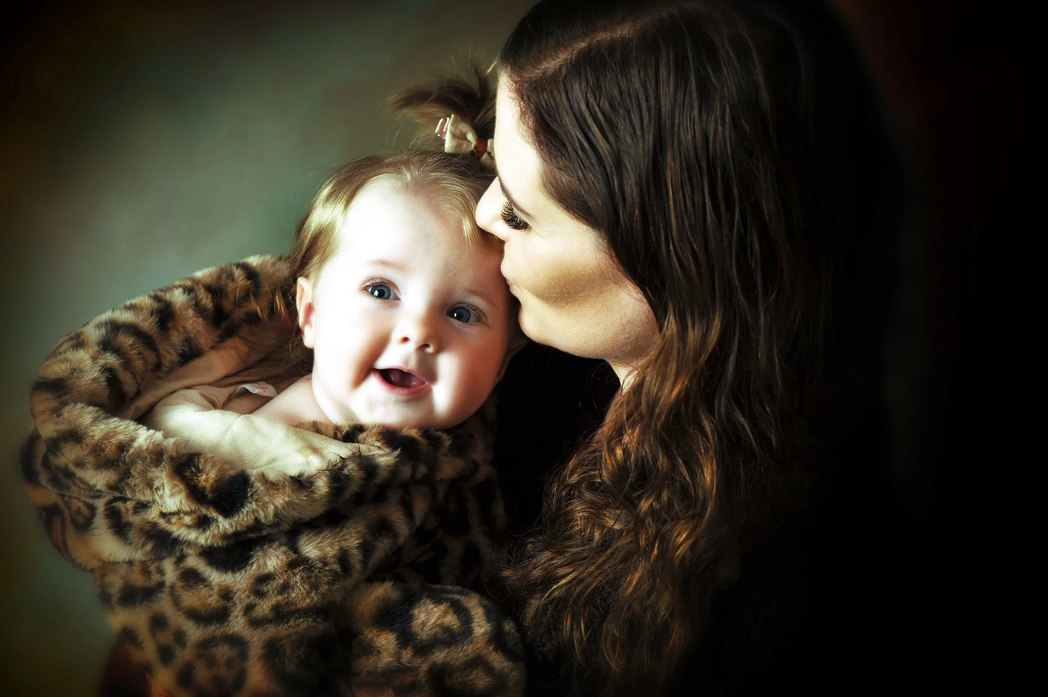 A woman with dark, wavy hair kissing a smiling child with blonde hair and blue eyes, wearing a leopard print coat.