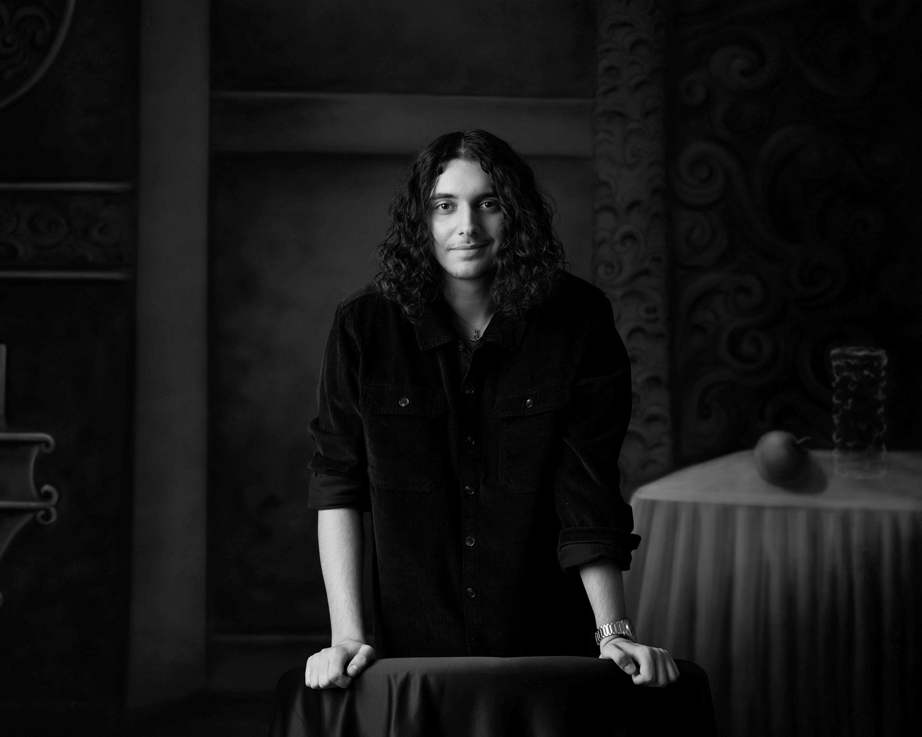Black and white portrait of a young man with shoulder-length curly hair, wearing a dark button-up shirt, standing with arms resting on a table, in front of a dark, textured background with decorative patterns.