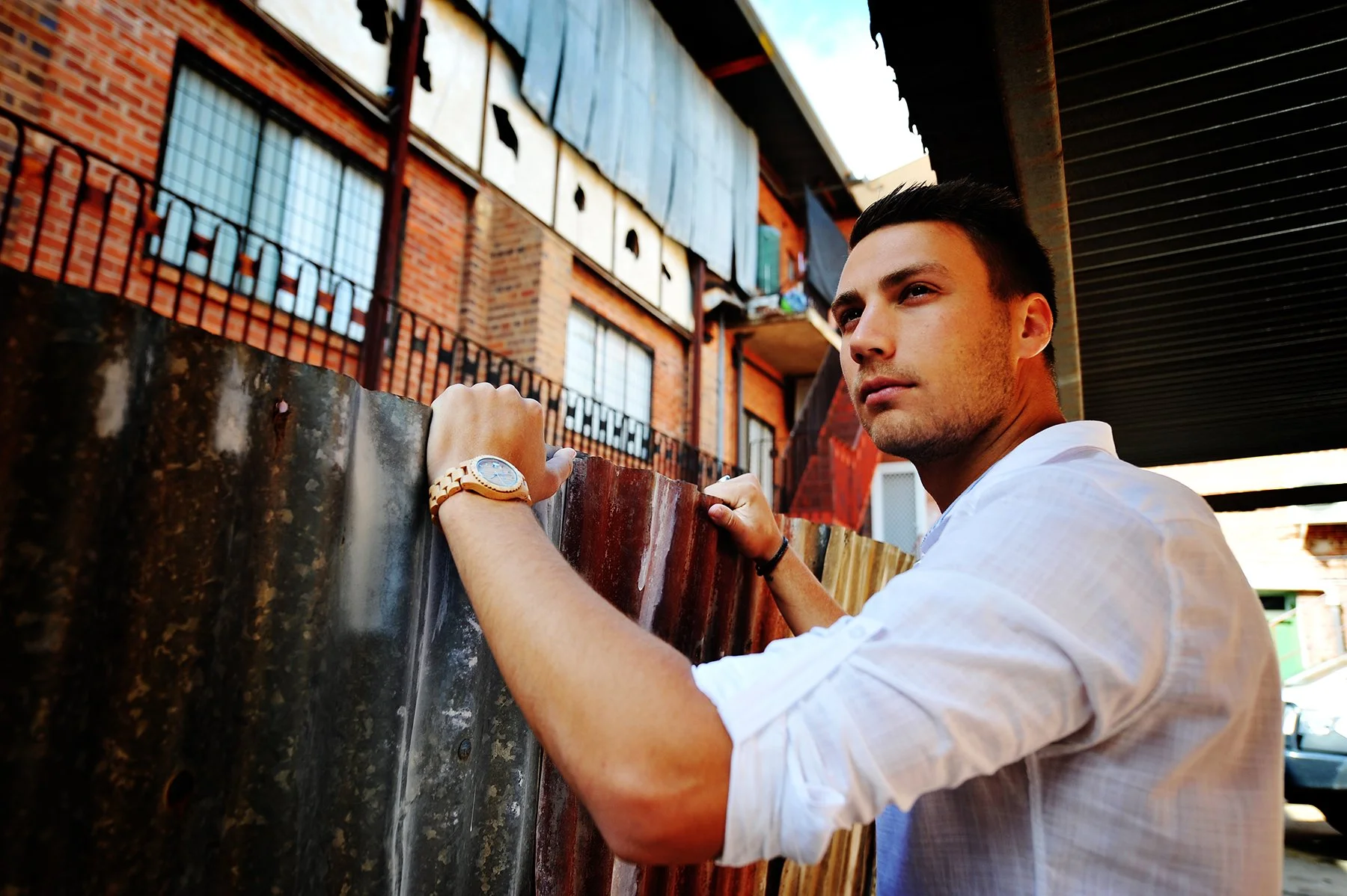 A young man with short dark hair wearing a white shirt and a gold wristwatch is leaning on a colorful metal fence, looking into the distance with a contemplative expression, in an urban setting with red brick buildings and laundry hanging above.