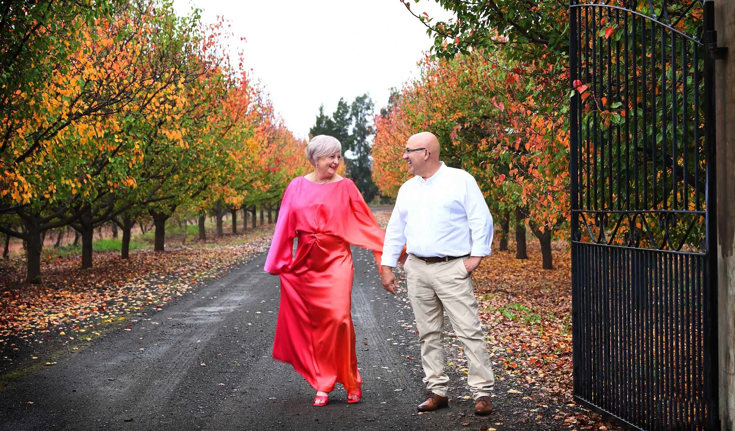 An elderly woman in a pink dress and a man in a white shirt walking and smiling together on a pathway surrounded by colorful fall trees.