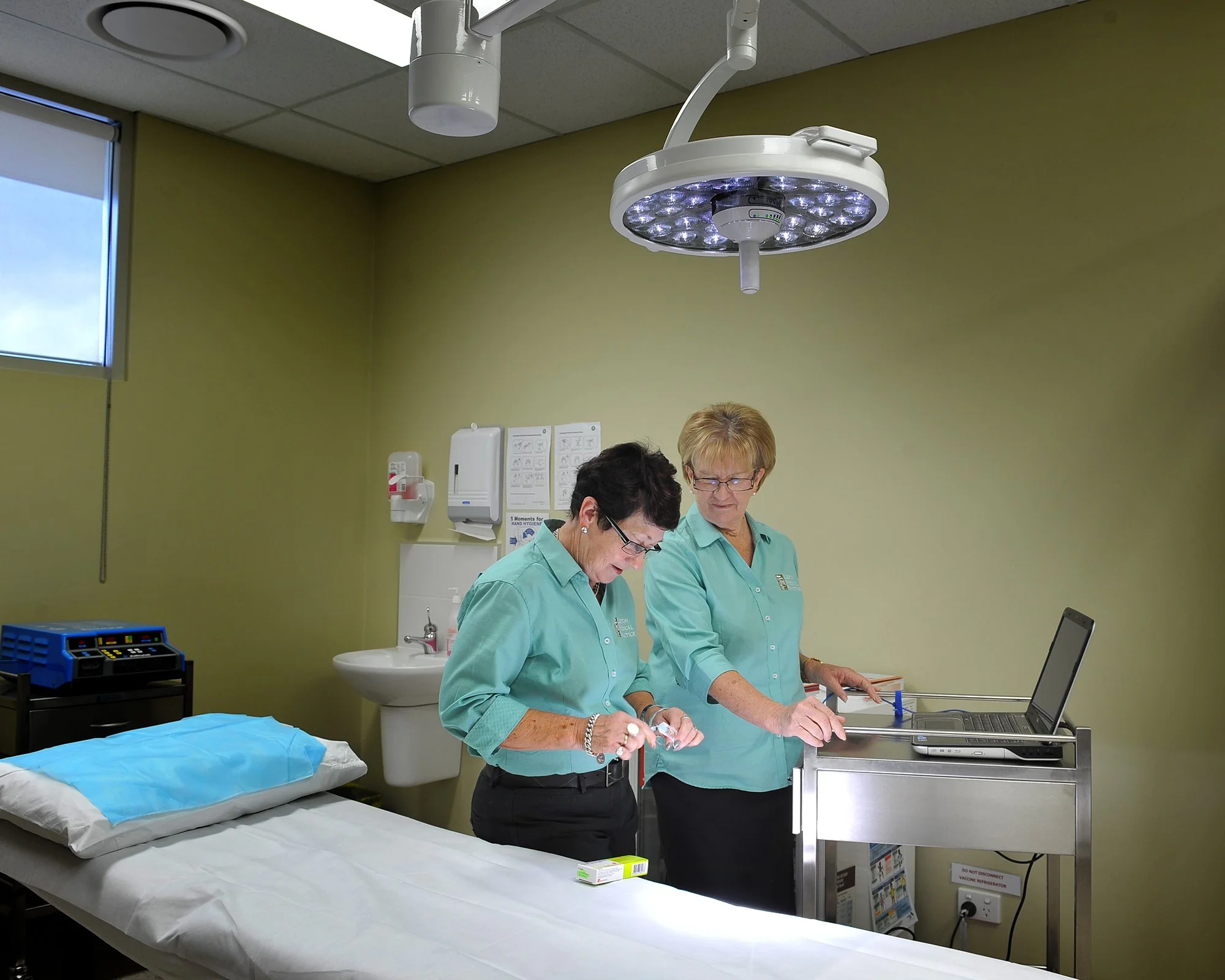 Two healthcare professionals in teal scrubs working in a medical examination room with a patient bed, medical equipment, and an overhead surgical light.