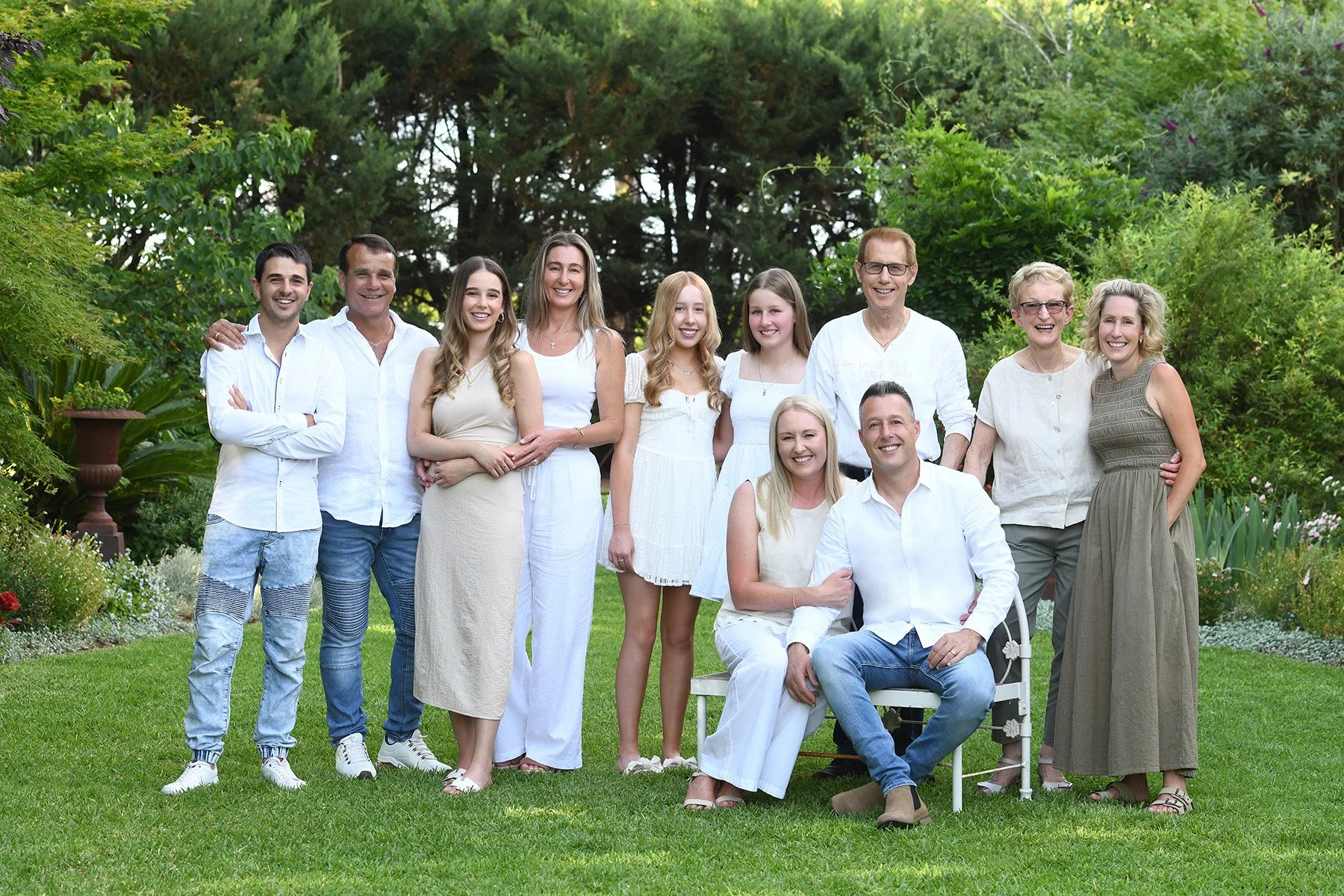 A large multi-generational family group posing outdoors in a lush green garden. The group includes men, women, and children, all dressed in light-colored summer clothing, smiling and standing close together.