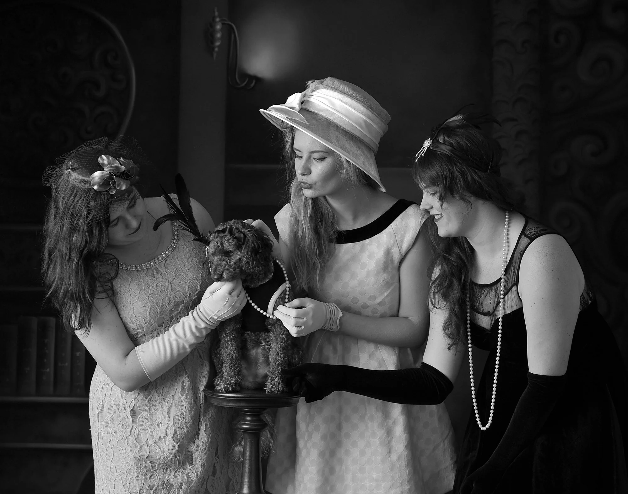Four women in vintage attire and hats are gathered around a table, holding a small dog dressed in clothing, examining or admiring it in a formal setting.