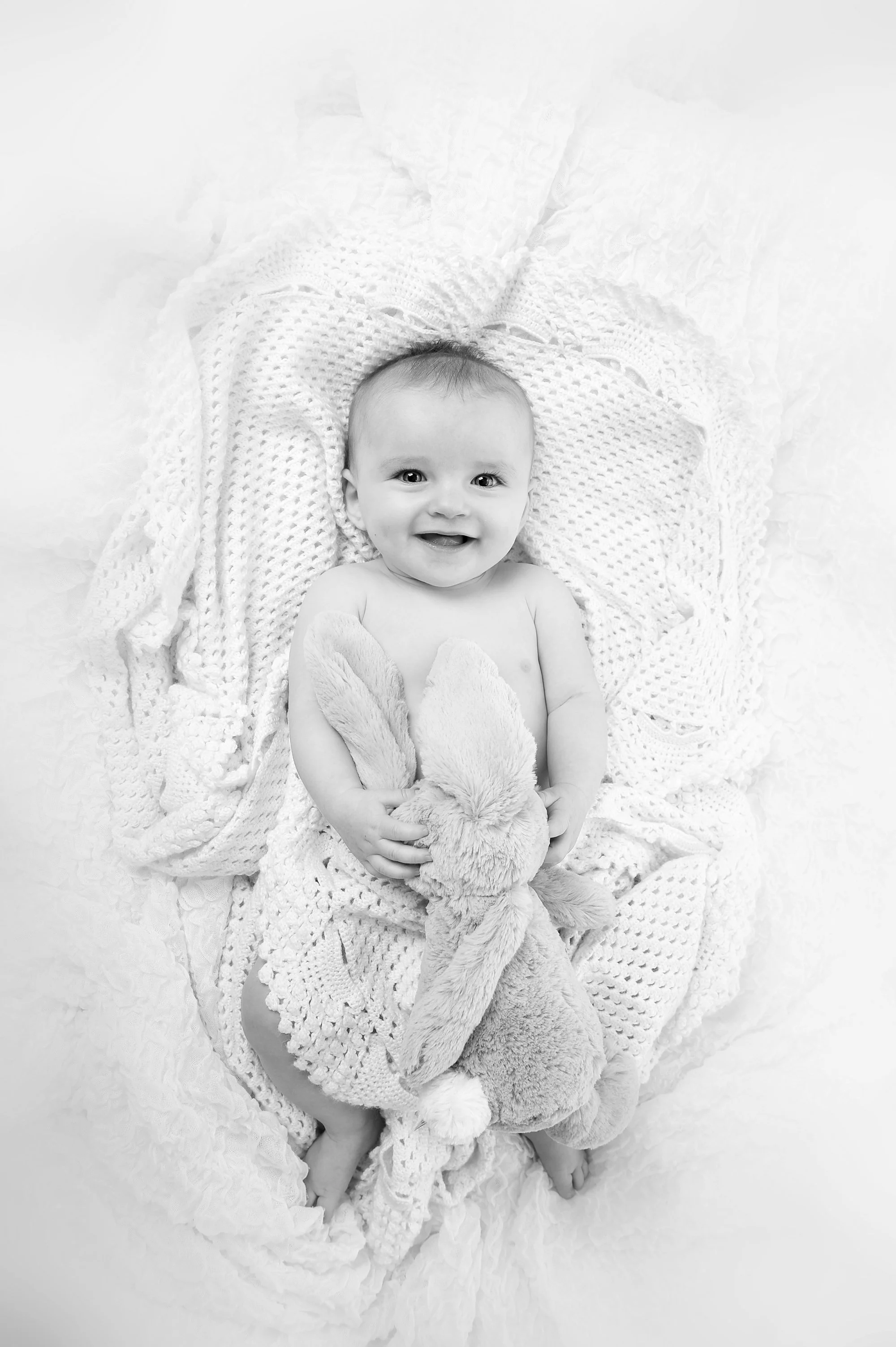 A baby with a wide smile, lying on a textured blanket, holding a soft bunny plush toy.