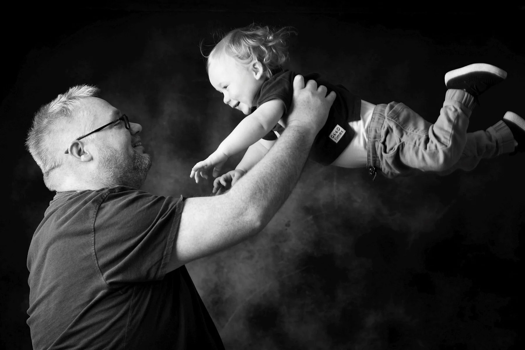 A black-and-white photo of a man lifting a young child in the air, both smiling happily, against a dark background.