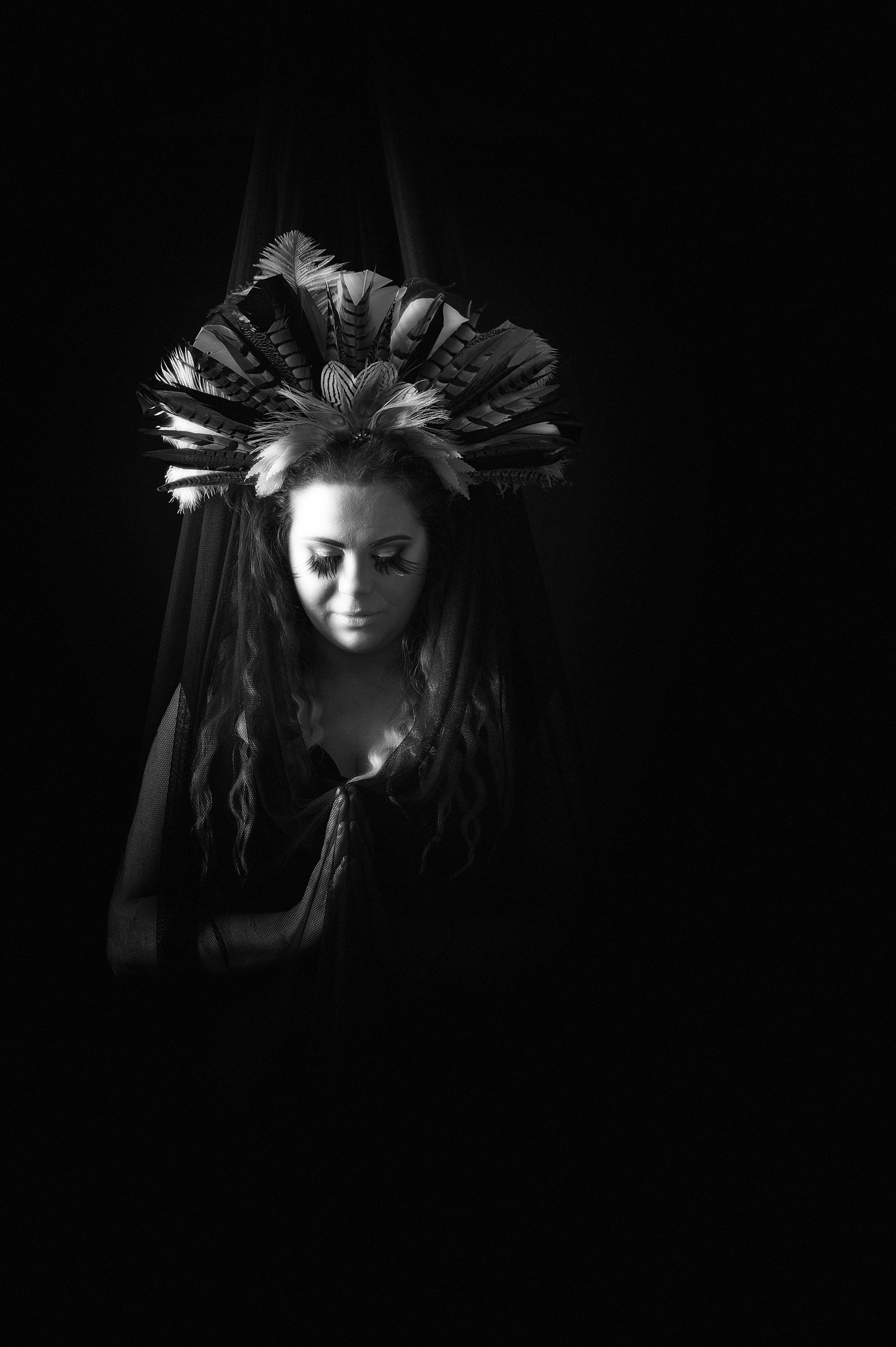 A woman with long hair and dramatic makeup, wearing an elaborate feathered headdress, standing against a black background, with her eyes closed and a veil draped over her shoulders.