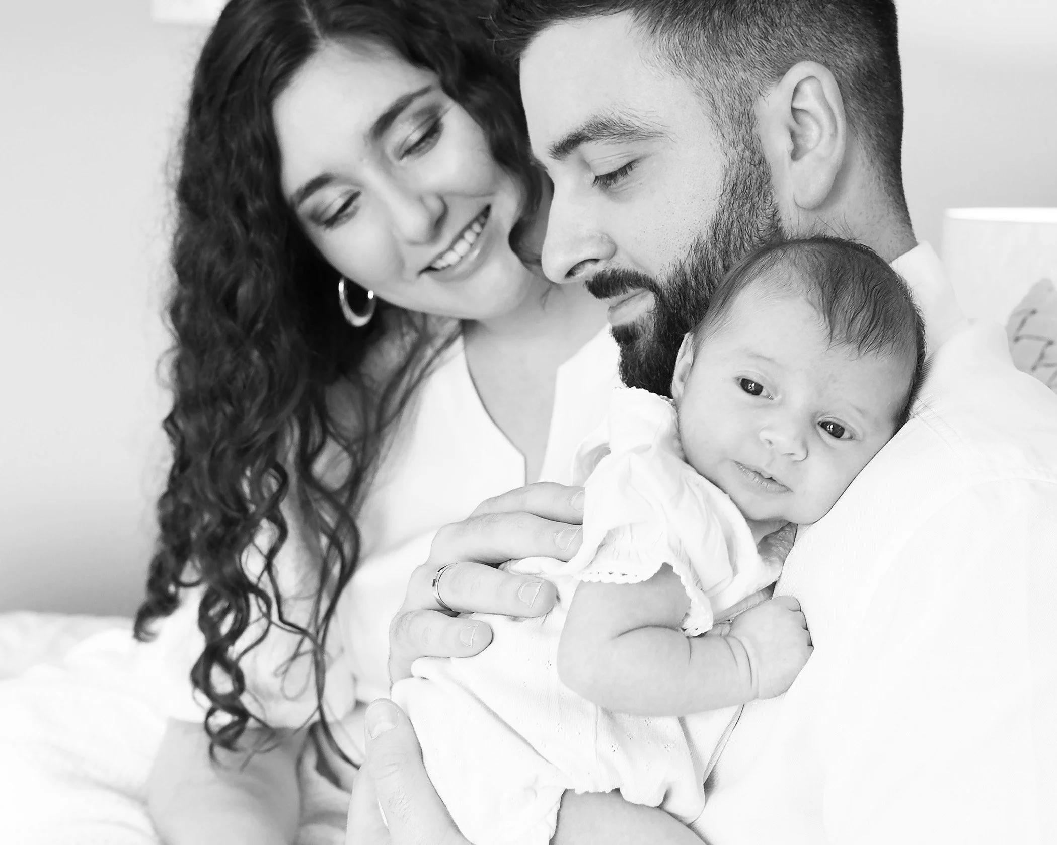 A black and white photo of a smiling woman with curly hair holding a baby, with a man close by, all appearing happy.