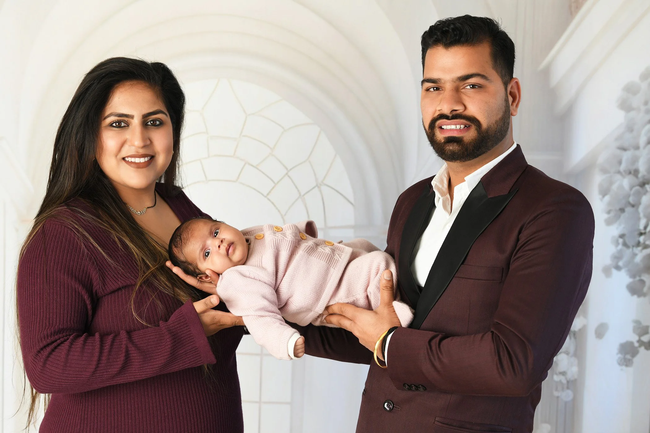 A family portrait of a woman with long dark hair, a man with short dark hair and a beard, and a baby in the woman's arms, against a light background with an arch and foliage.
