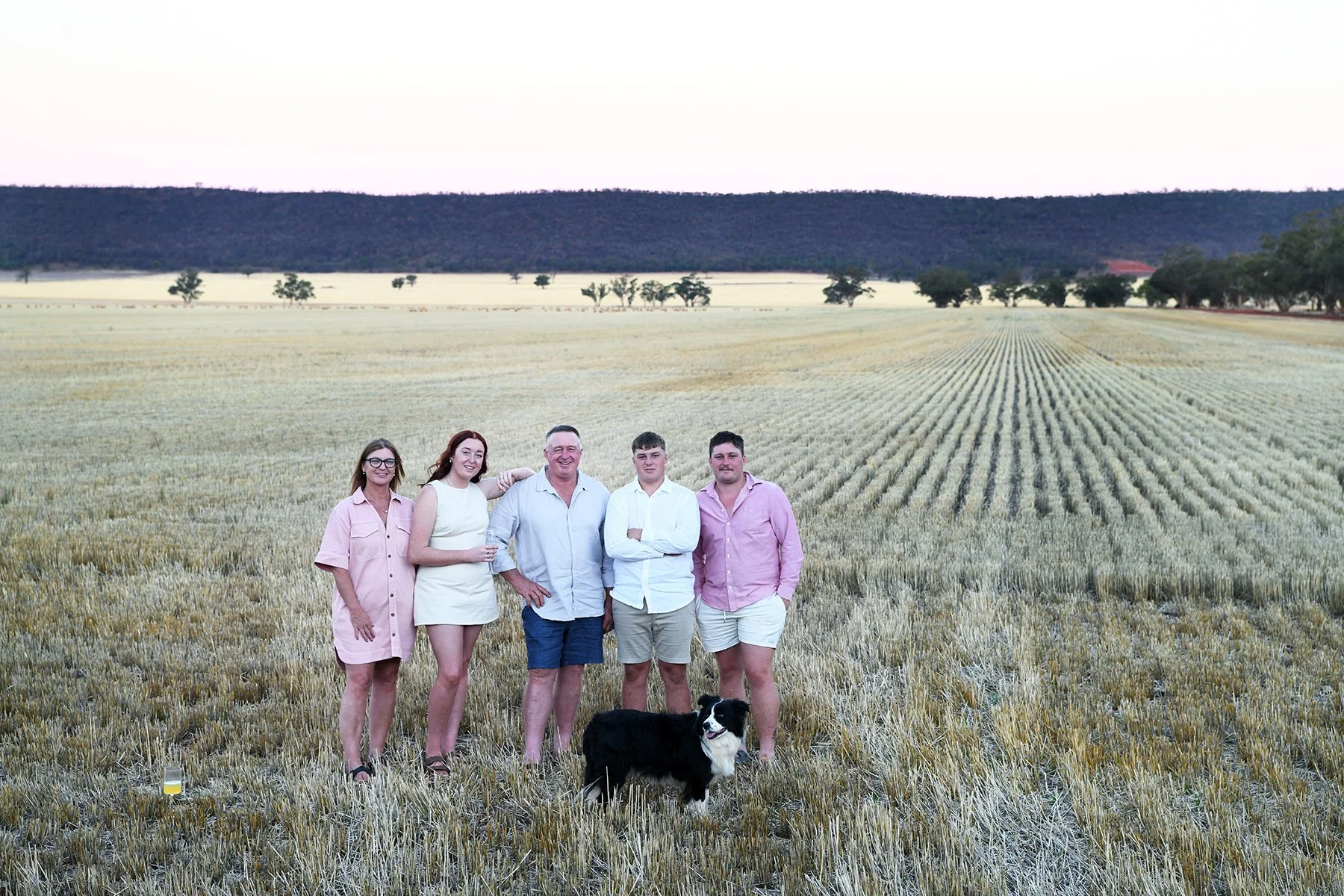 Group of five people standing in a wheat field with a black and white dog in front. The background features a landscape with trees and distant hills under a clear sky.