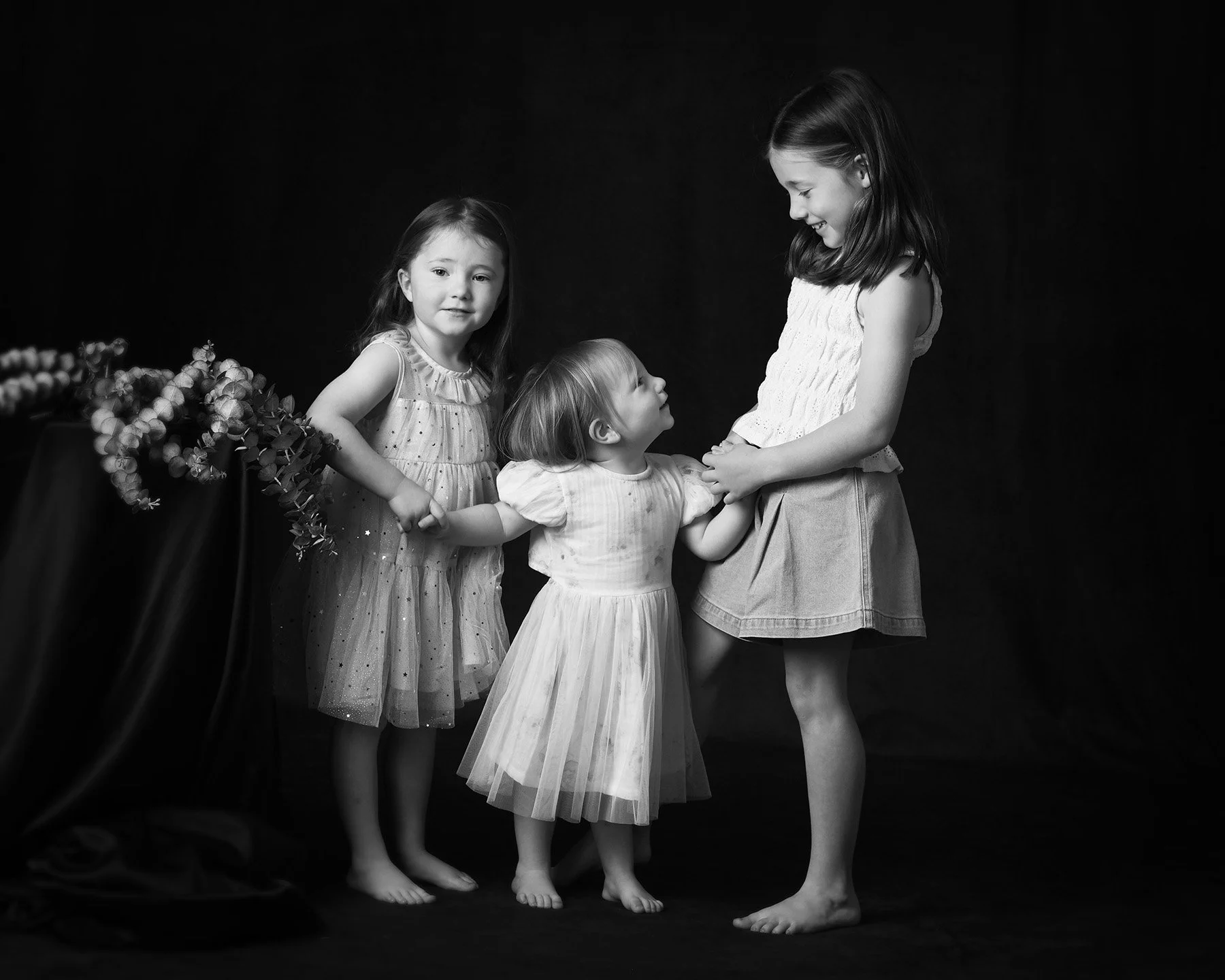 Four girls, three young and one older, wearing light-colored dresses, standing together on a dark background. The young girl in the center looks up at the older girl, who is smiling and looking down at her. The girl on the left stands next to a table