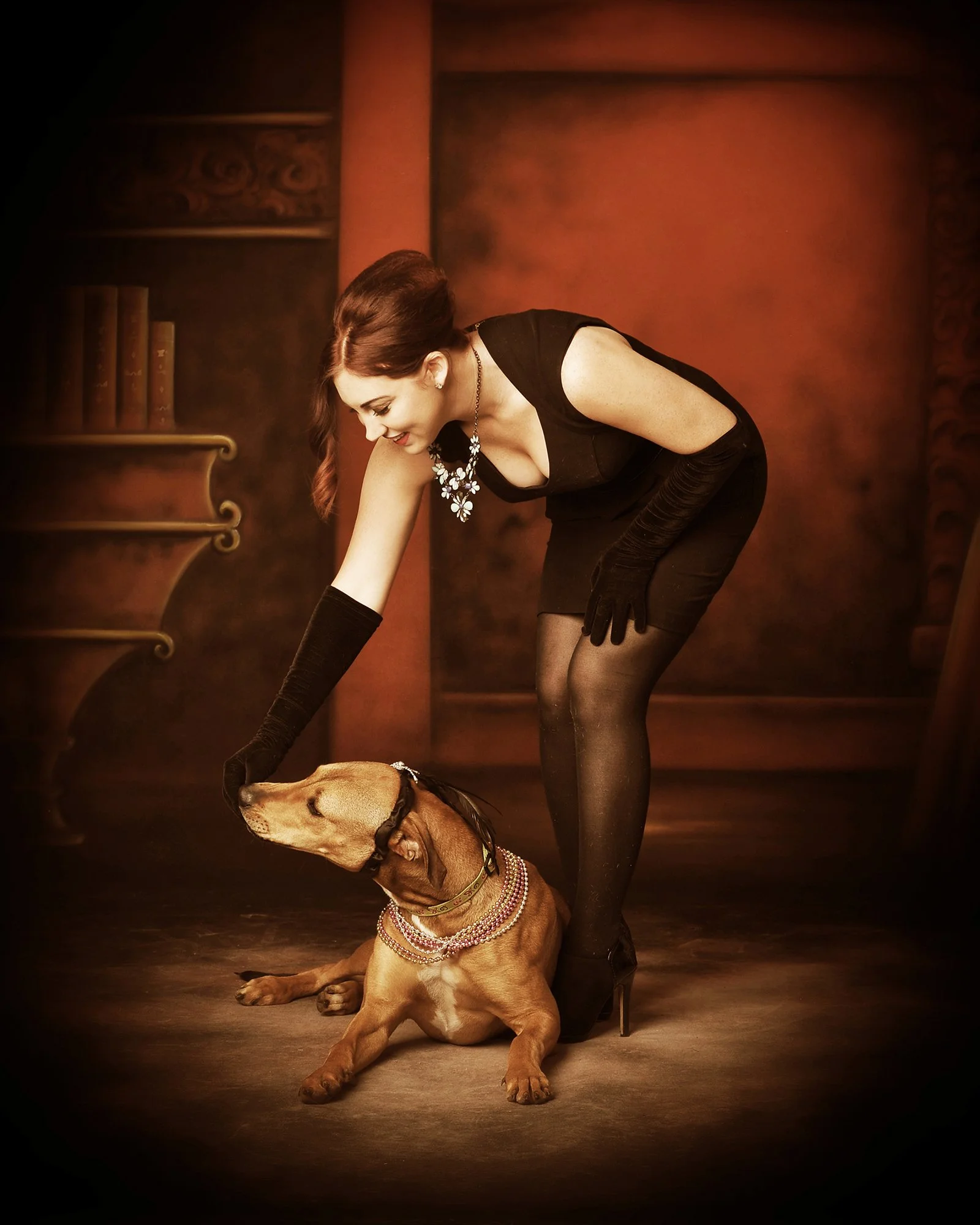 A woman in black dress and long gloves bending over a sitting brown dog adorned with pearl necklaces, set against a reddish-brown background with ornate wooden shelves.