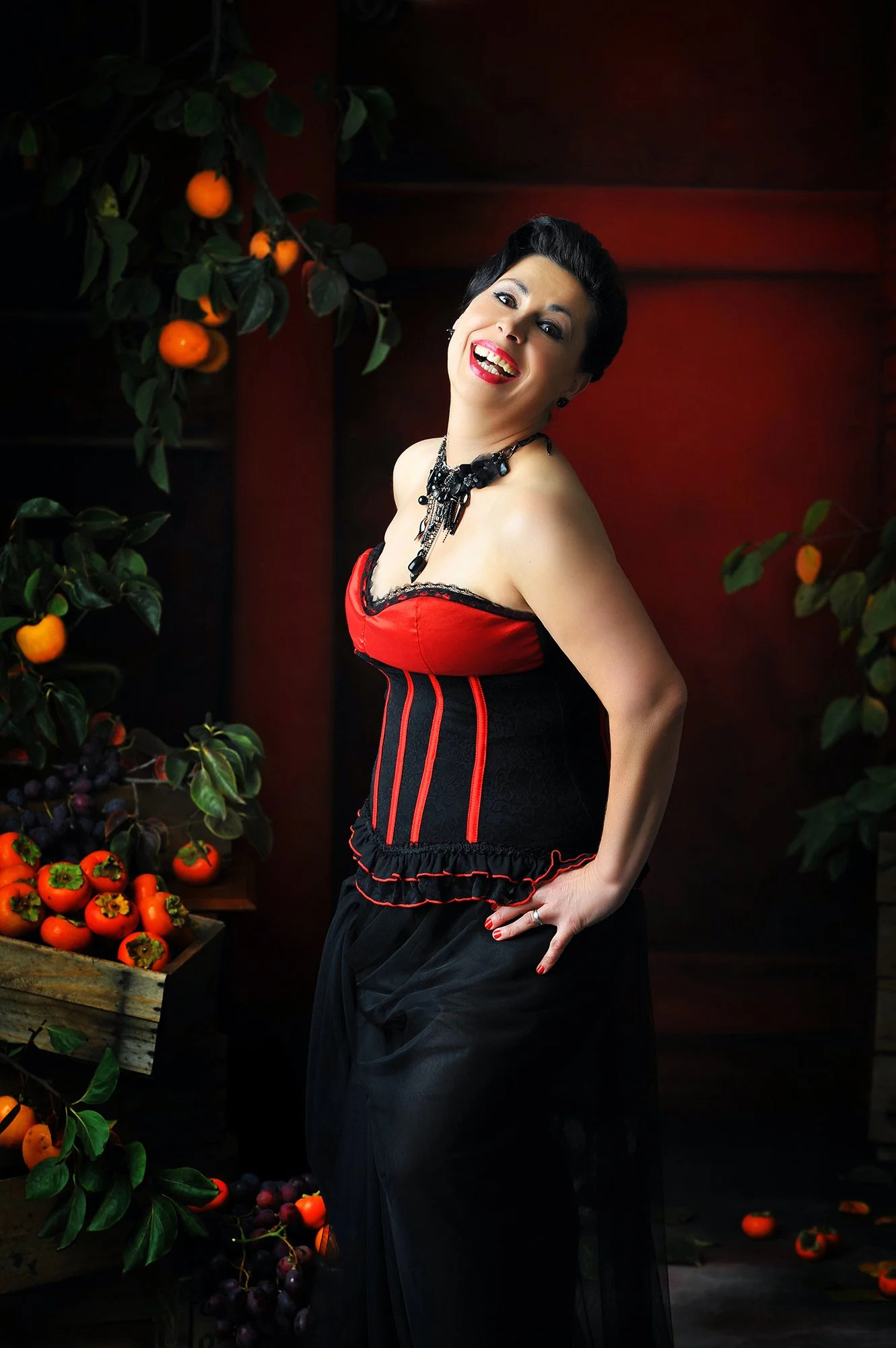 A woman with short dark hair and bright red lipstick, wearing a black and red corset-style dress and a black statement necklace, stands in front of a dark wood background surrounded by hanging persimmon and grape vines, with some persimmons and grape