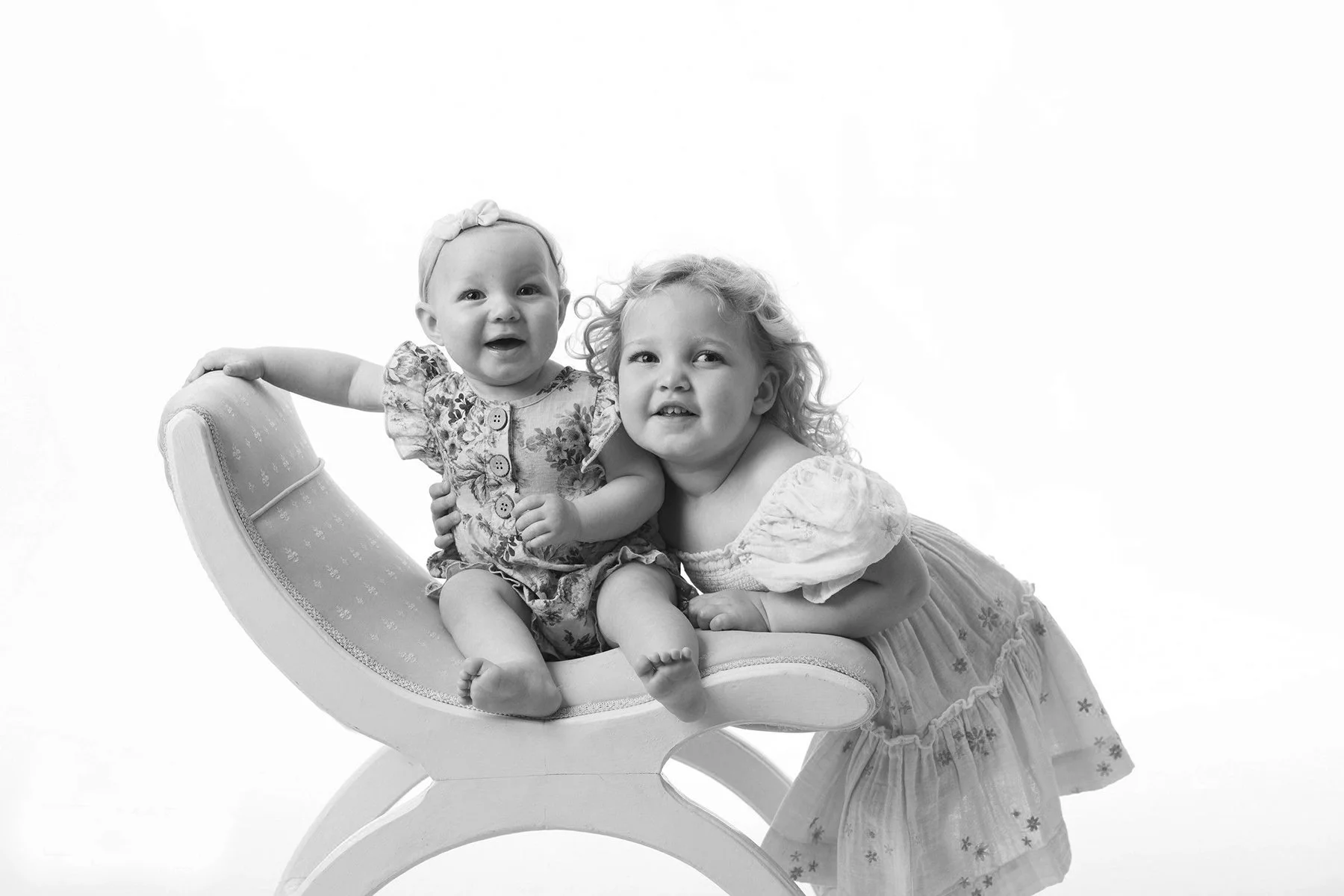 Two young girls, one sitting on a vintage chaise lounge and the other leaning on her, both smiling and looking at the camera. The image is in black and white.