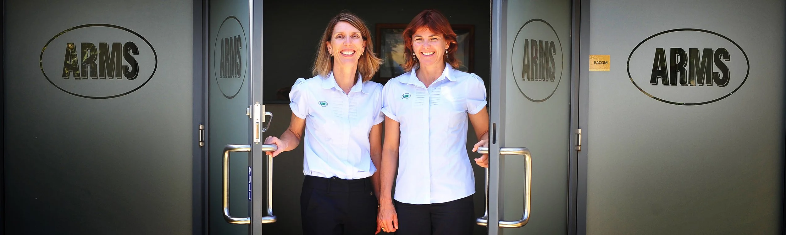 Two women smiling and standing at the entrance of a building with glass doors that has 'ARMS' written on it, wearing white shirts with ARMS logos.