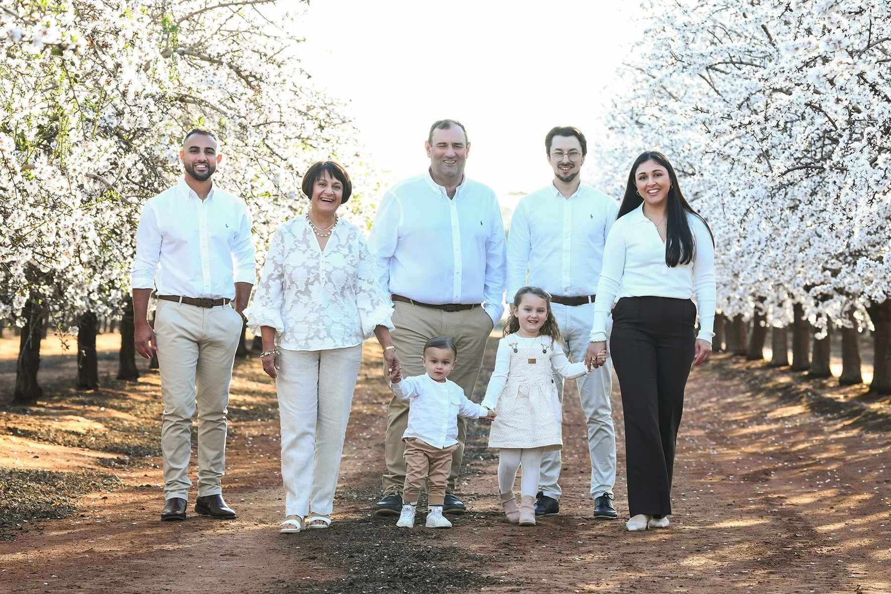 A multigenerational family walking together through a blooming orchard of white flowers, dressed in white and light-colored clothing, holding hands and smiling.