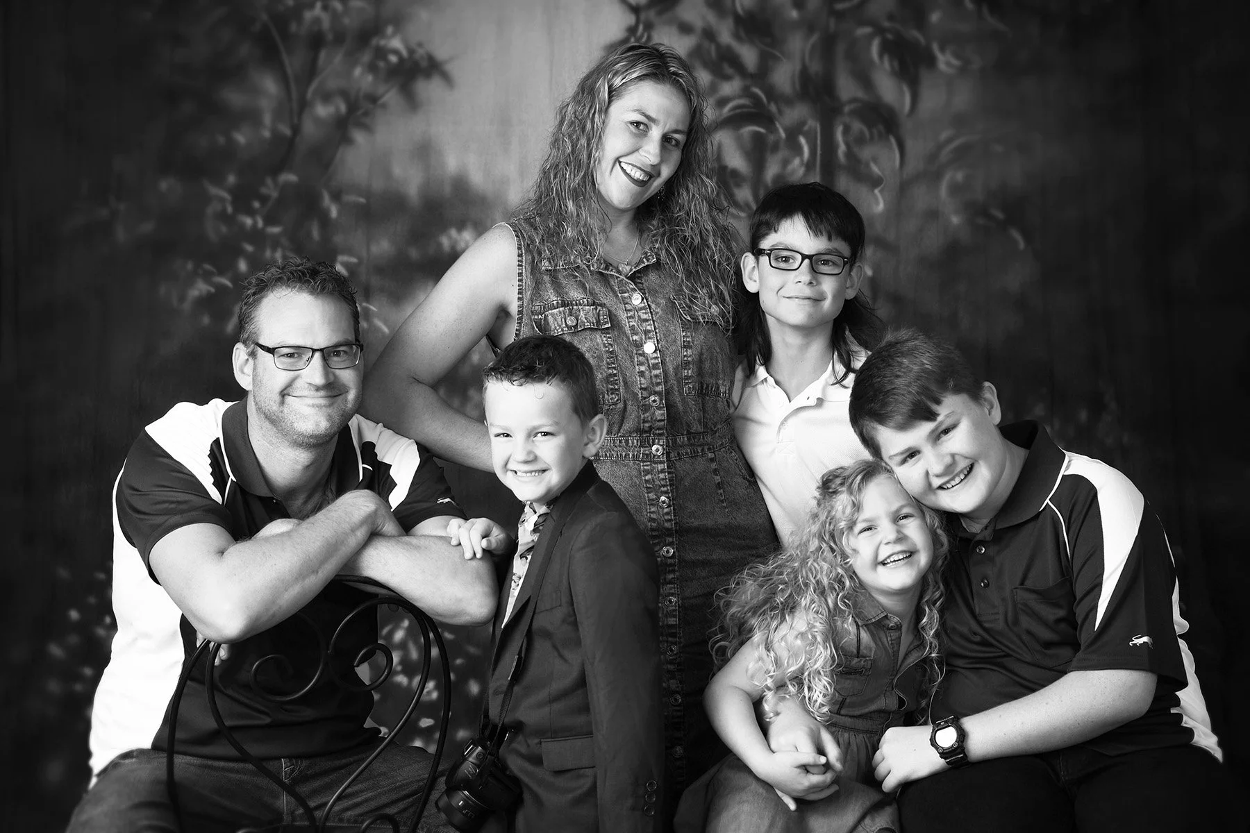 A black and white family portrait of seven members, including a woman, four boys, and two girls, posing together smiling against a blurred outdoor background.