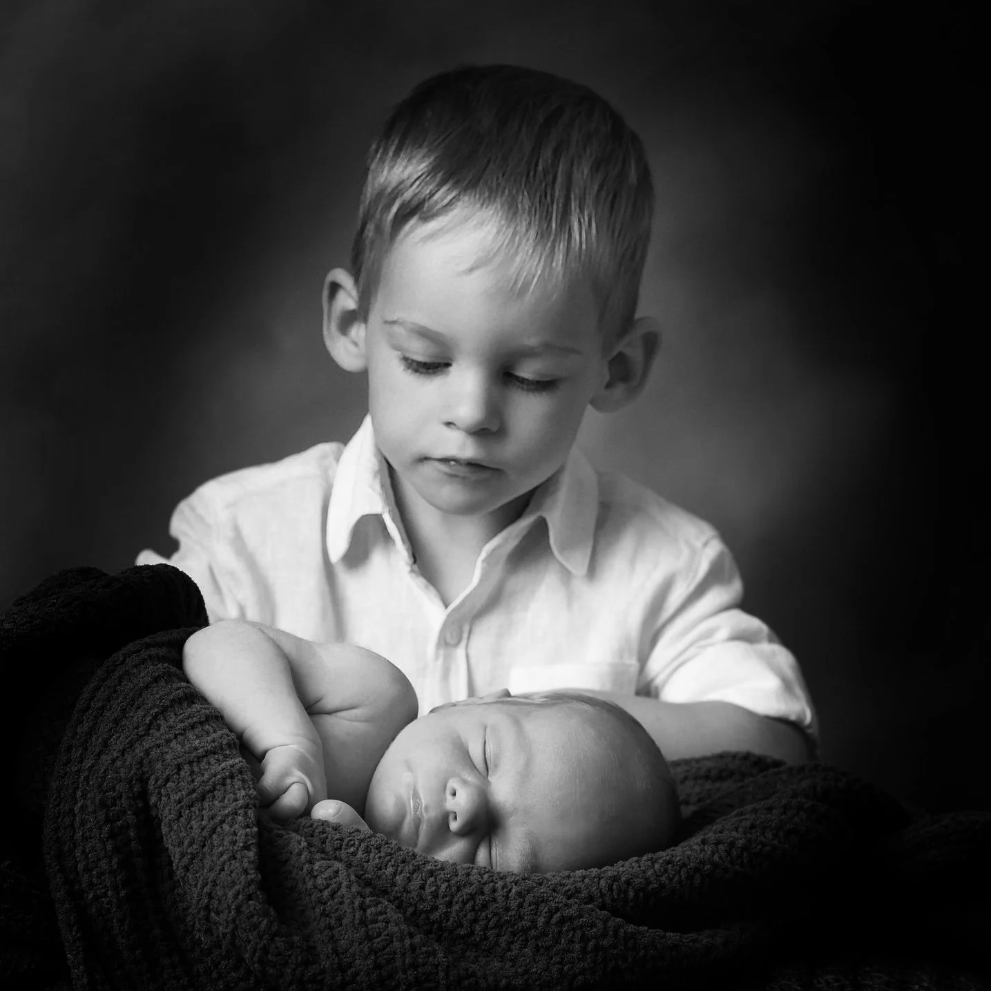A young boy with short hair gazes at a sleeping newborn baby wrapped in a blanket.