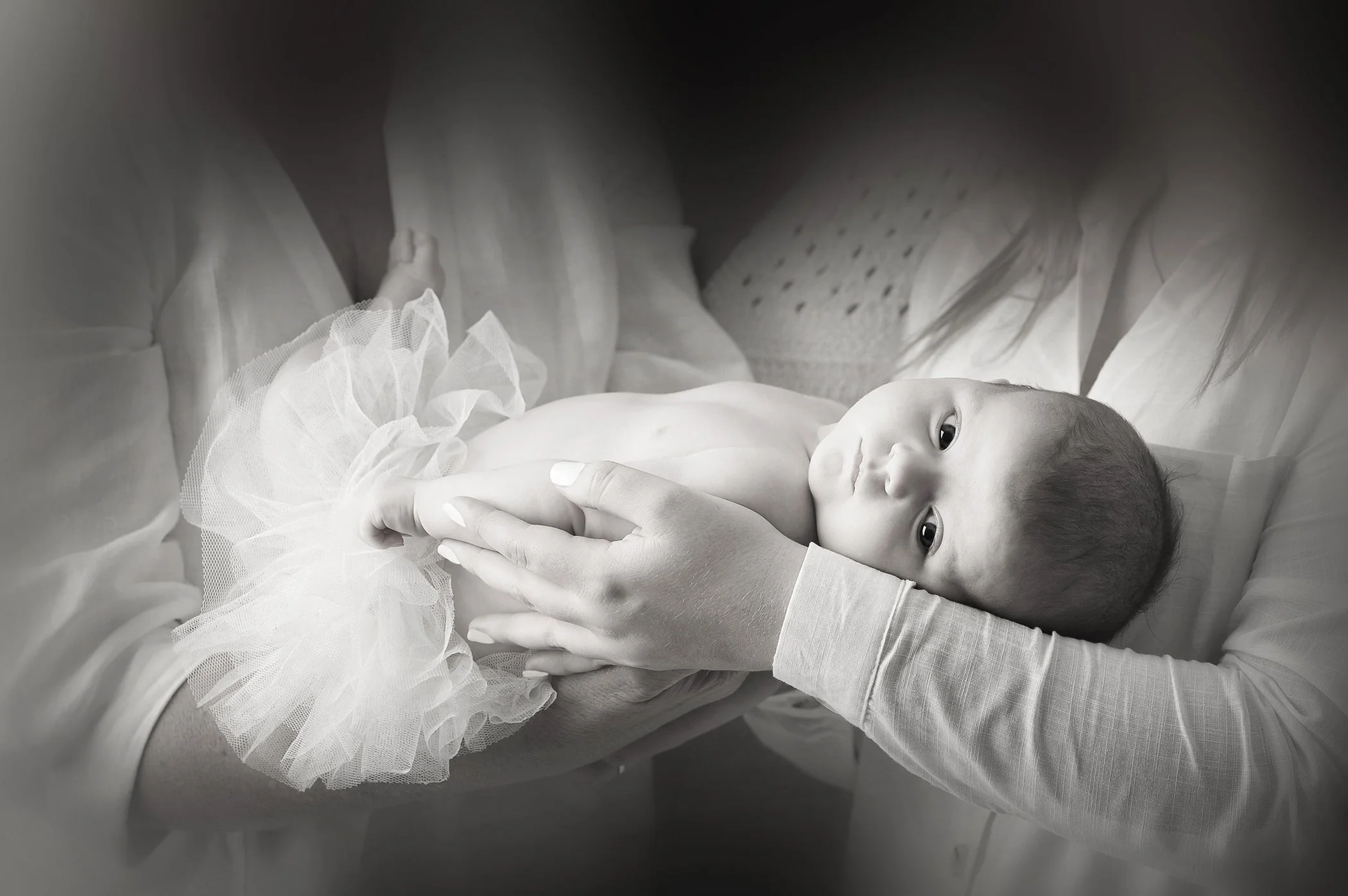 A newborn baby lying on a hospital bed, being cradled by a caregiver's hands, with the baby wrapped in a white, lacy blanket. The baby has wide open eyes, and the scene is in black and white.