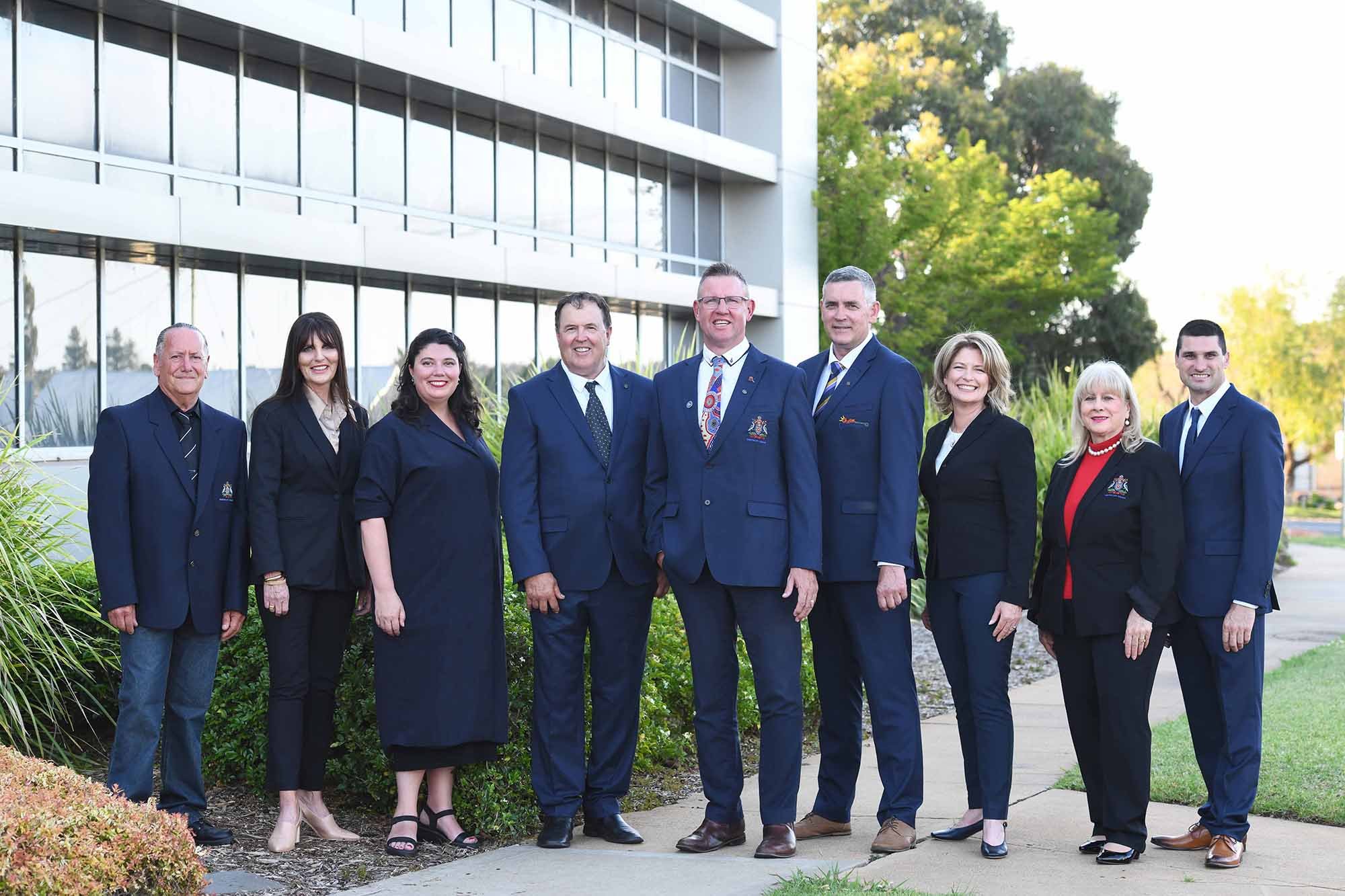 A group of nine professionally dressed people standing outside in front of a modern office building with large glass windows and green trees. The group includes men and women smiling at the camera.