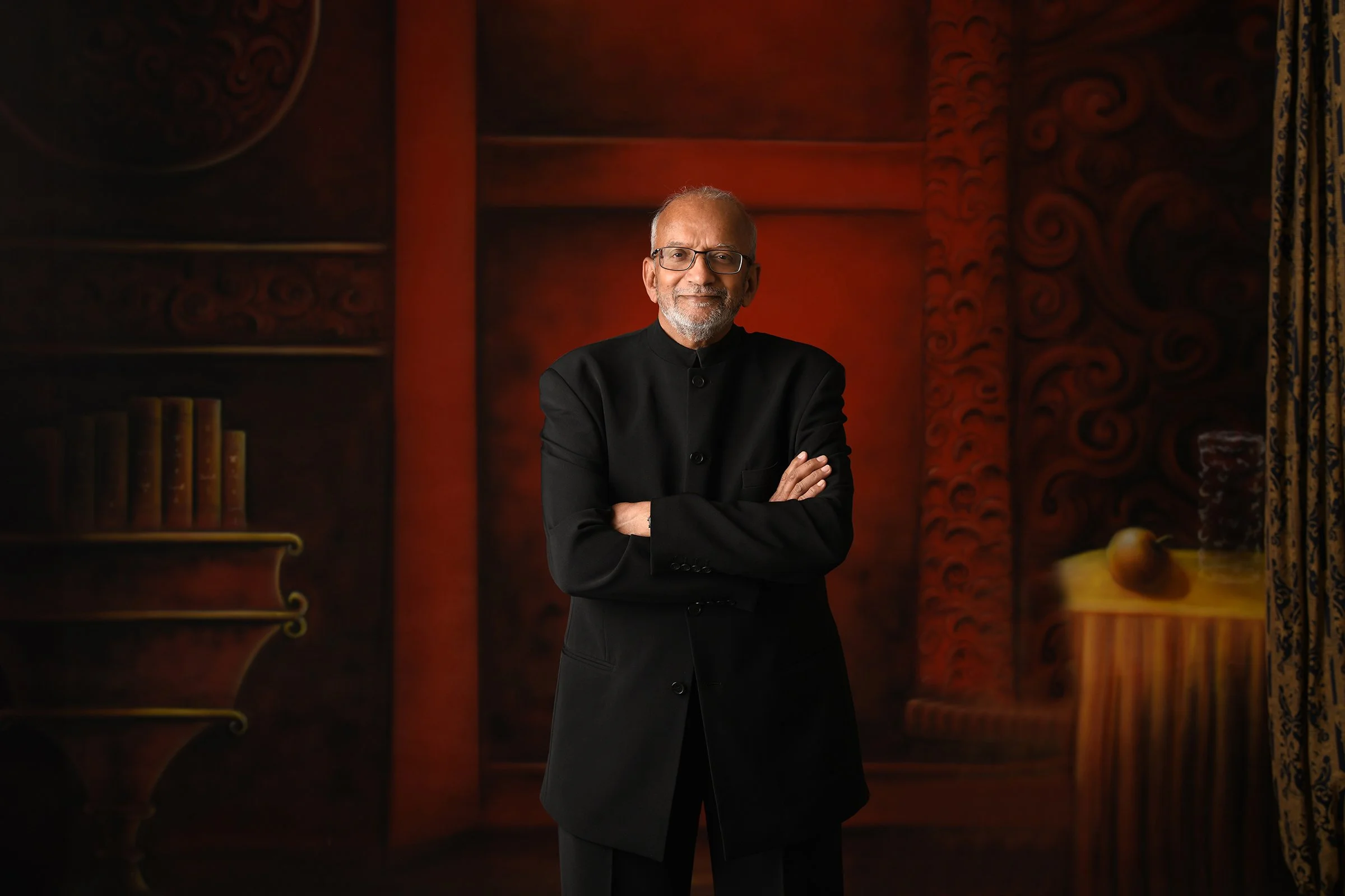 A man with glasses and a beard wearing a black suit, standing with arms crossed in front of a painted background that resembles a classical interior with shelves, books, and decorative curtains.