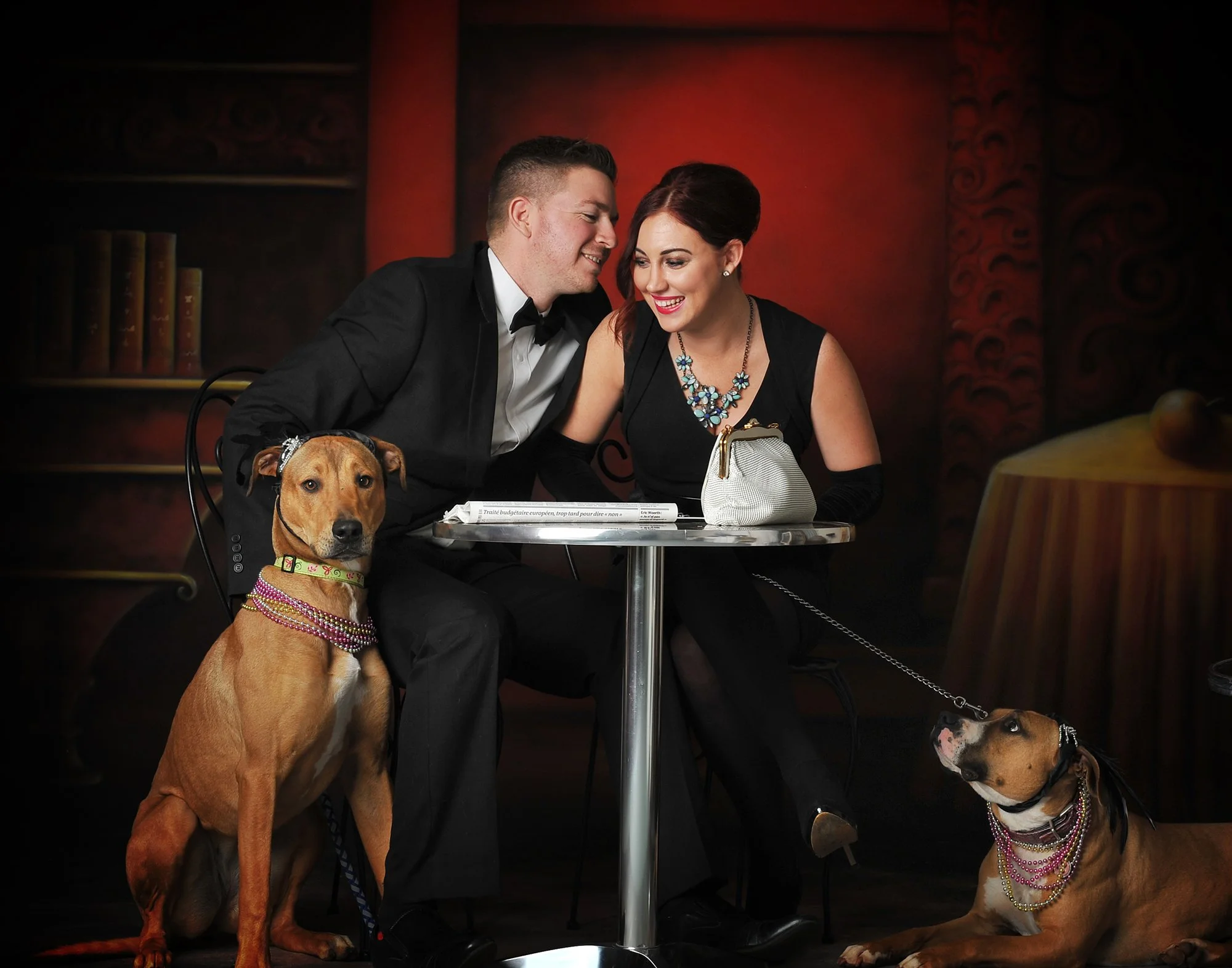 A man and woman sitting at a table, dressed in elegant formal attire, with two dogs adorned in jewelry, in a warmly lit restaurant with dark decor.