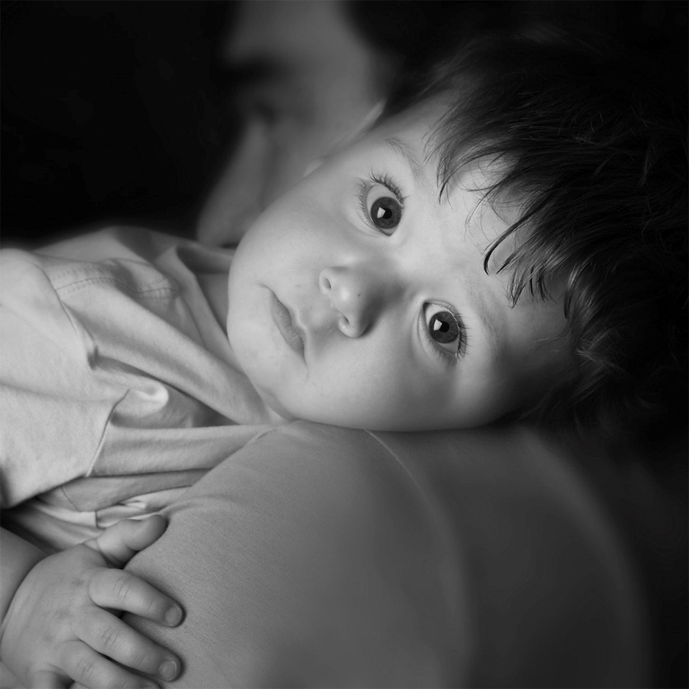 A young child with big eyes and tousled hair looking thoughtfully to the side, resting on an adult's shoulder in a black and white photograph.