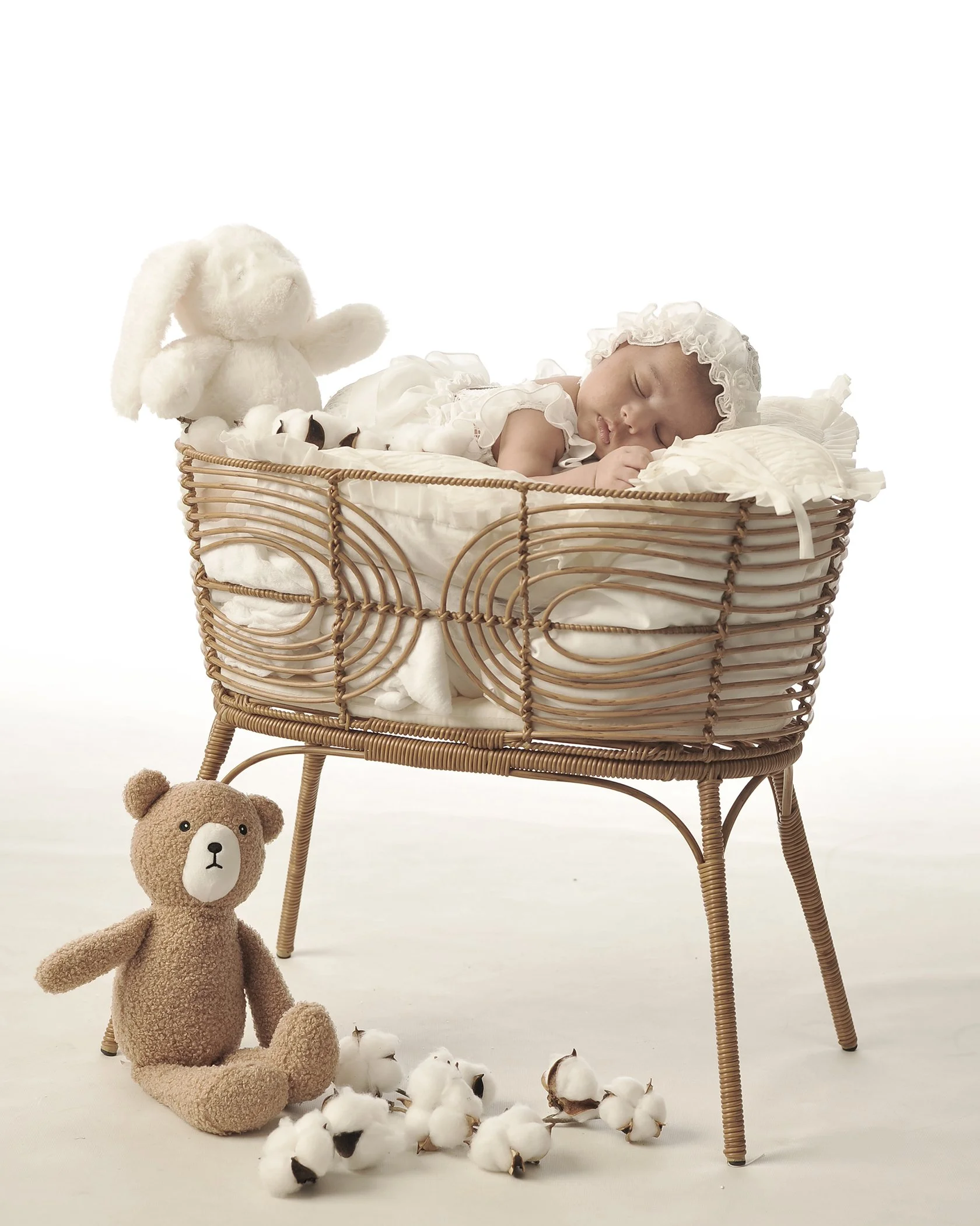 A sleeping baby wearing a white outfit and bonnet, lying in a wicker basket with white bedding. A plush bunny toy is sitting in the basket beside the baby. A beige teddy bear is sitting on the floor in front of the basket, surrounded by white cotton 